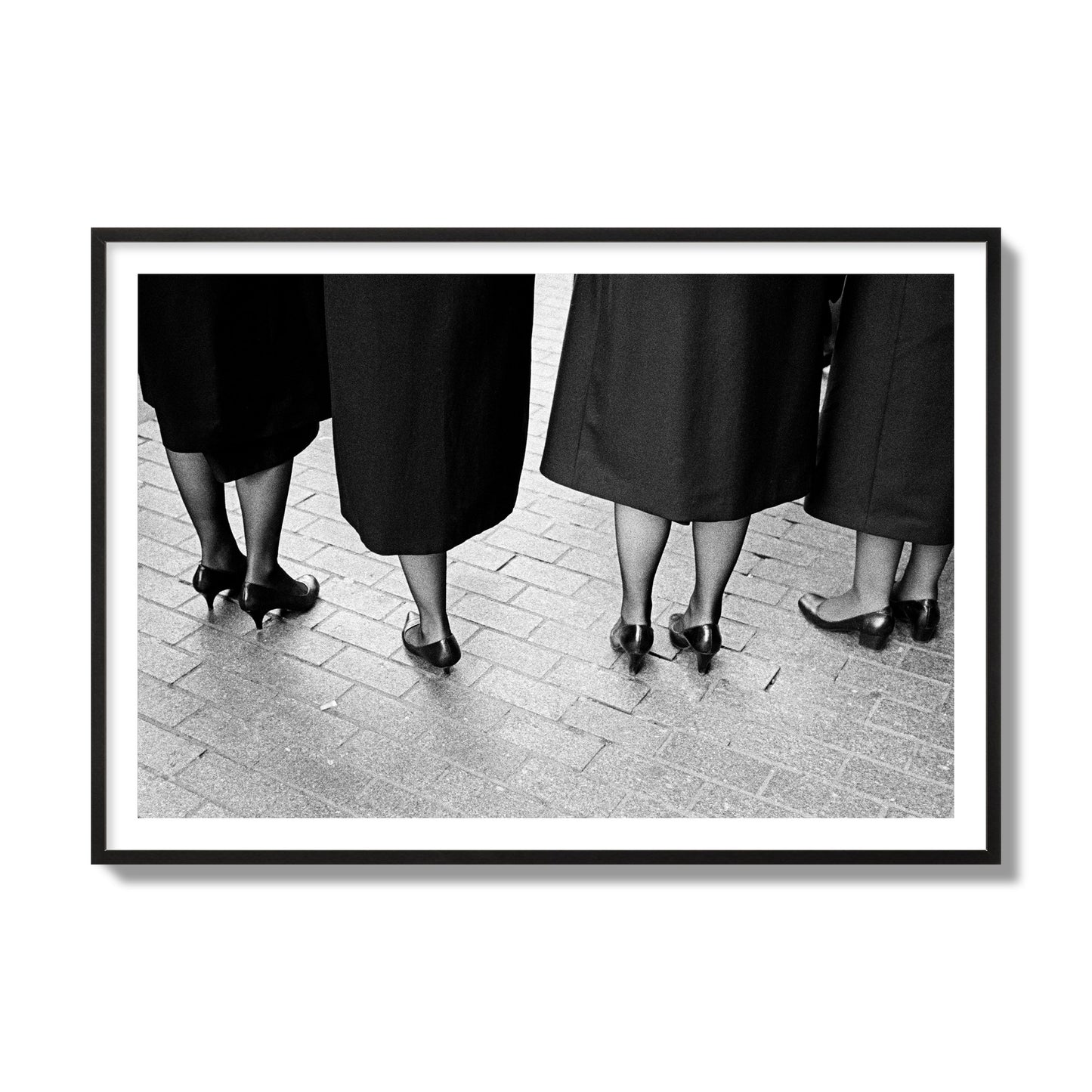 Fine art photograph depicting four women’s feet in high heels standing on a brick-paved street. One woman is notably missing a leg, adding a striking element to the composition, archival pigment print, collector size, black frame.