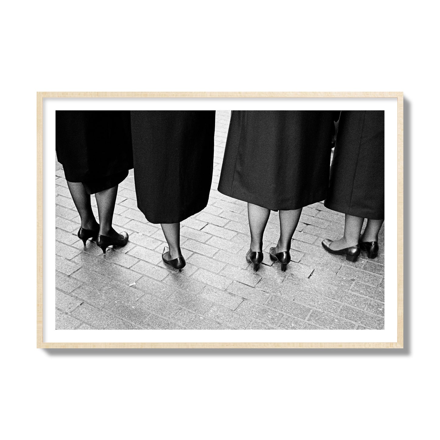 Fine art photograph depicting four women’s feet in high heels standing on a brick-paved street. One woman is notably missing a leg, adding a striking element to the composition, archival pigment print, collector size, natural wood frame.