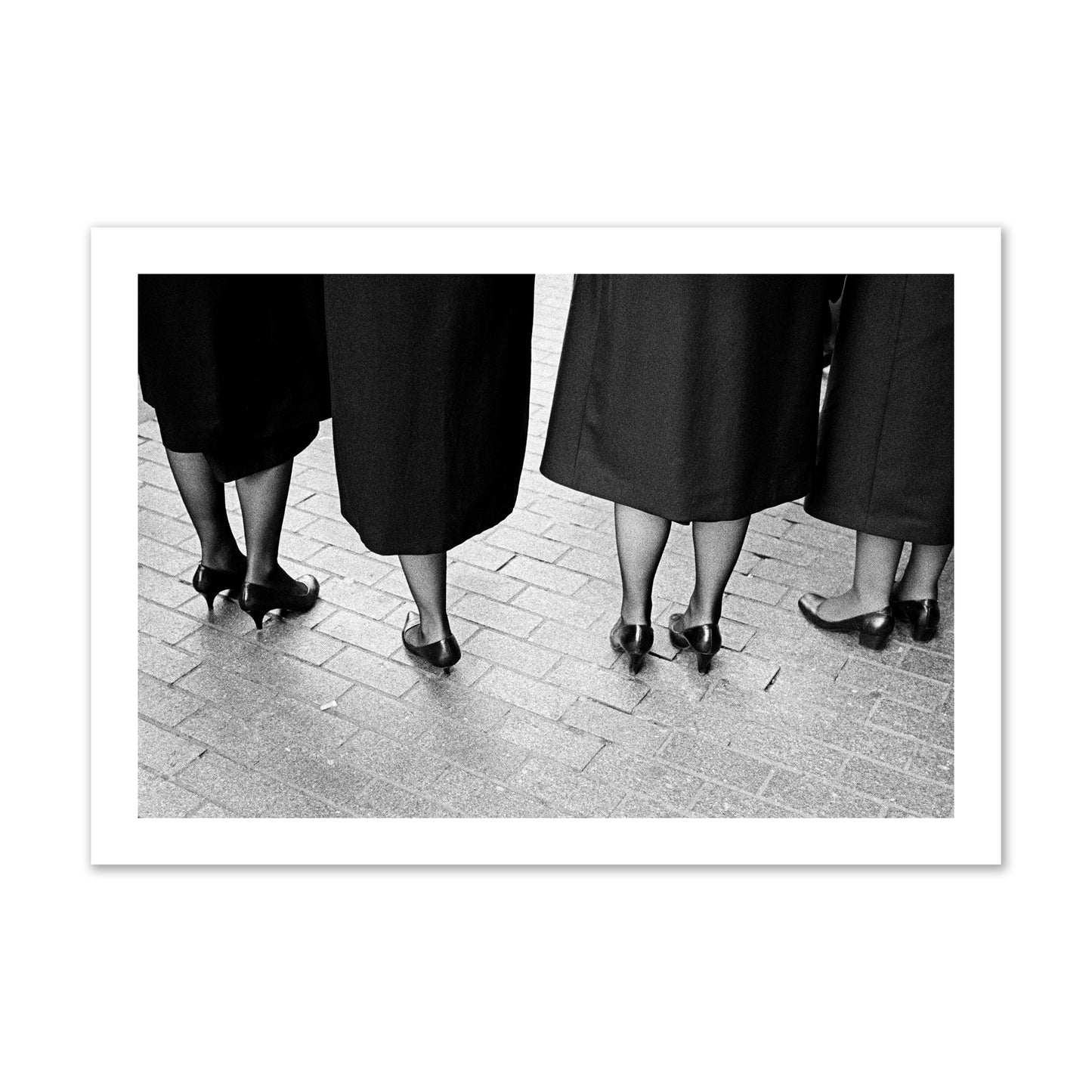 Fine art photograph depicting four women’s feet in high heels standing on a brick-paved street. One woman is notably missing a leg, adding a striking element to the composition, archival pigment print, collector size, unframed.