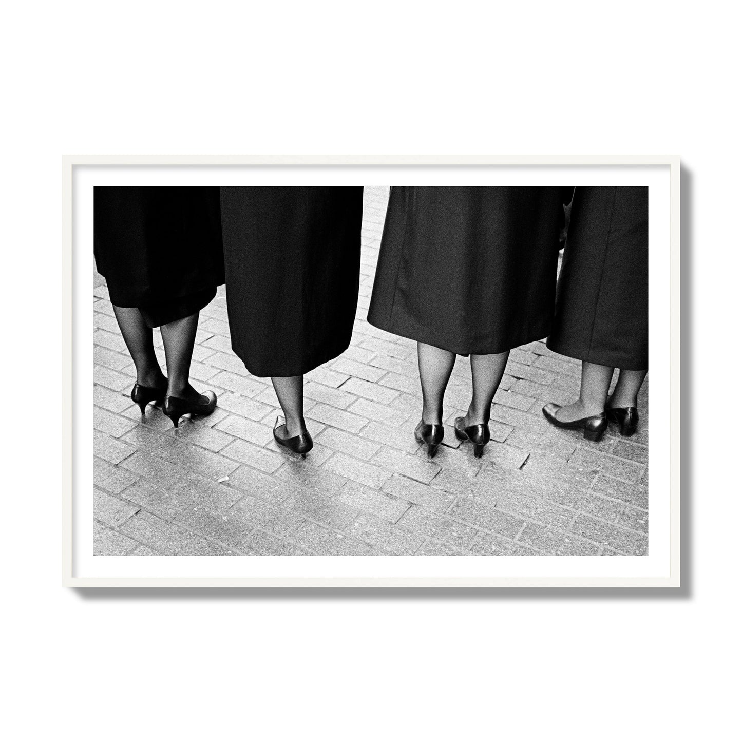 Fine art photograph depicting four women’s feet in high heels standing on a brick-paved street. One woman is notably missing a leg, adding a striking element to the composition, archival pigment print, collector size, white frame.