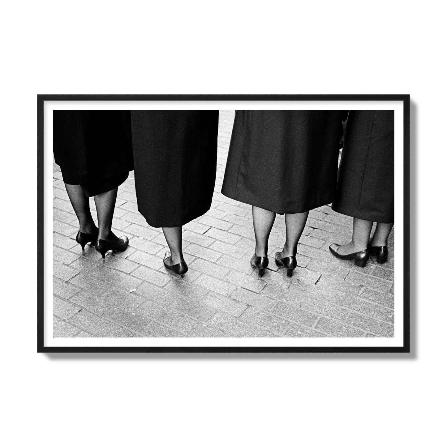 Fine art photograph depicting four women’s feet in high heels standing on a brick-paved street. One woman is notably missing a leg, adding a striking element to the composition, archival pigment print, gallery size, black frame.