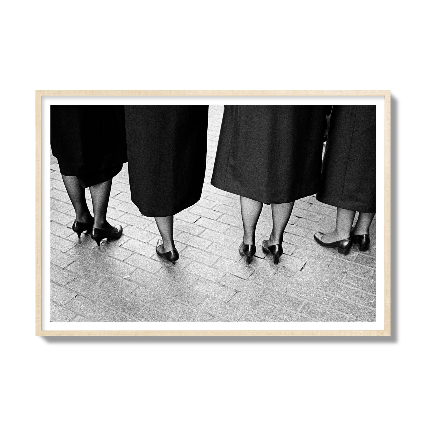 Fine art photograph depicting four women’s feet in high heels standing on a brick-paved street. One woman is notably missing a leg, adding a striking element to the composition, archival pigment print, gallery size, natural wood frame.