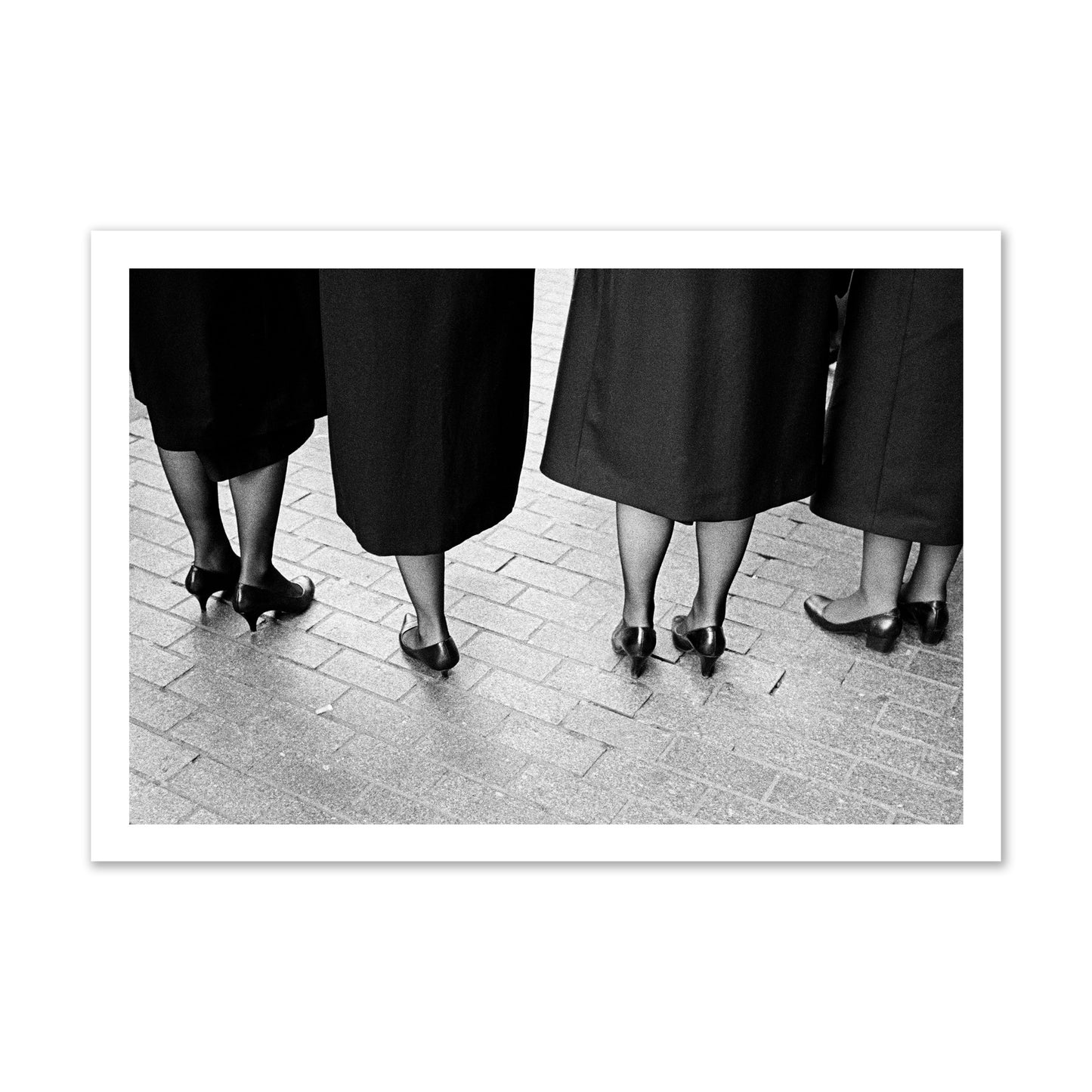 Fine art photograph depicting four women’s feet in high heels standing on a brick-paved street. One woman is notably missing a leg, adding a striking element to the composition, archival pigment print, gallery size, unframed.