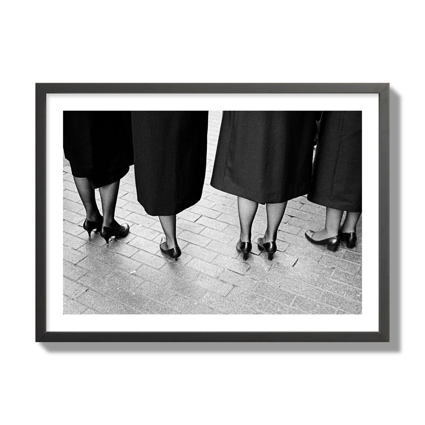 Fine art photograph depicting four women’s feet in high heels standing on a brick-paved street. One woman is notably missing a leg, adding a striking element to the composition, archival pigment print, giant size, black frame.