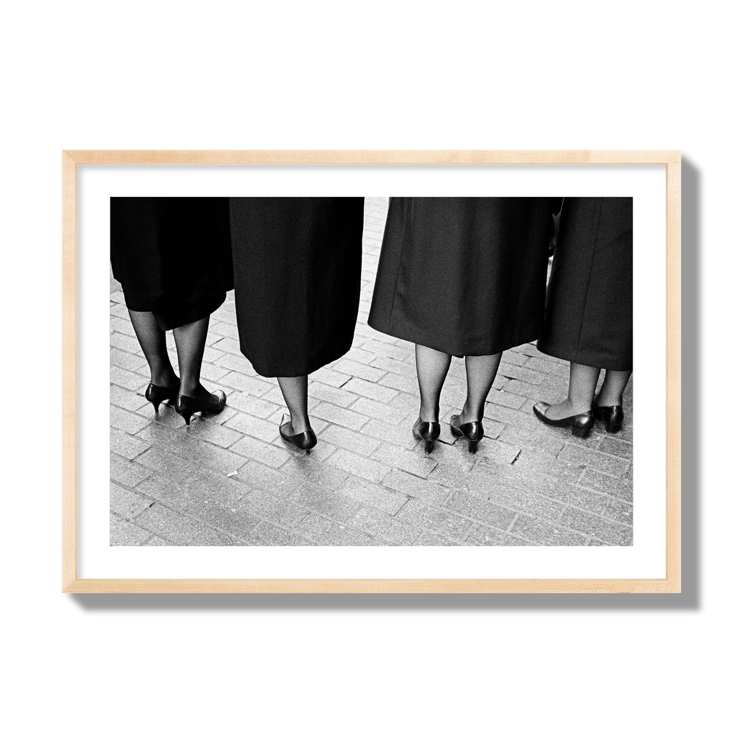 Fine art photograph depicting four women’s feet in high heels standing on a brick-paved street. One woman is notably missing a leg, adding a striking element to the composition, archival pigment print, giant size, natural wood frame.