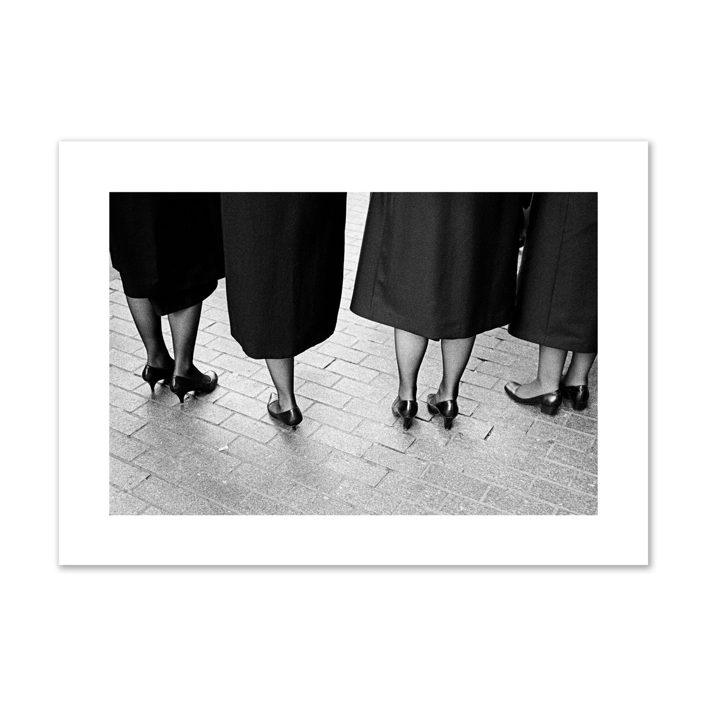 Fine art photograph depicting four women’s feet in high heels standing on a brick-paved street. One woman is notably missing a leg, adding a striking element to the composition, archival pigment print, giant size, unframed.