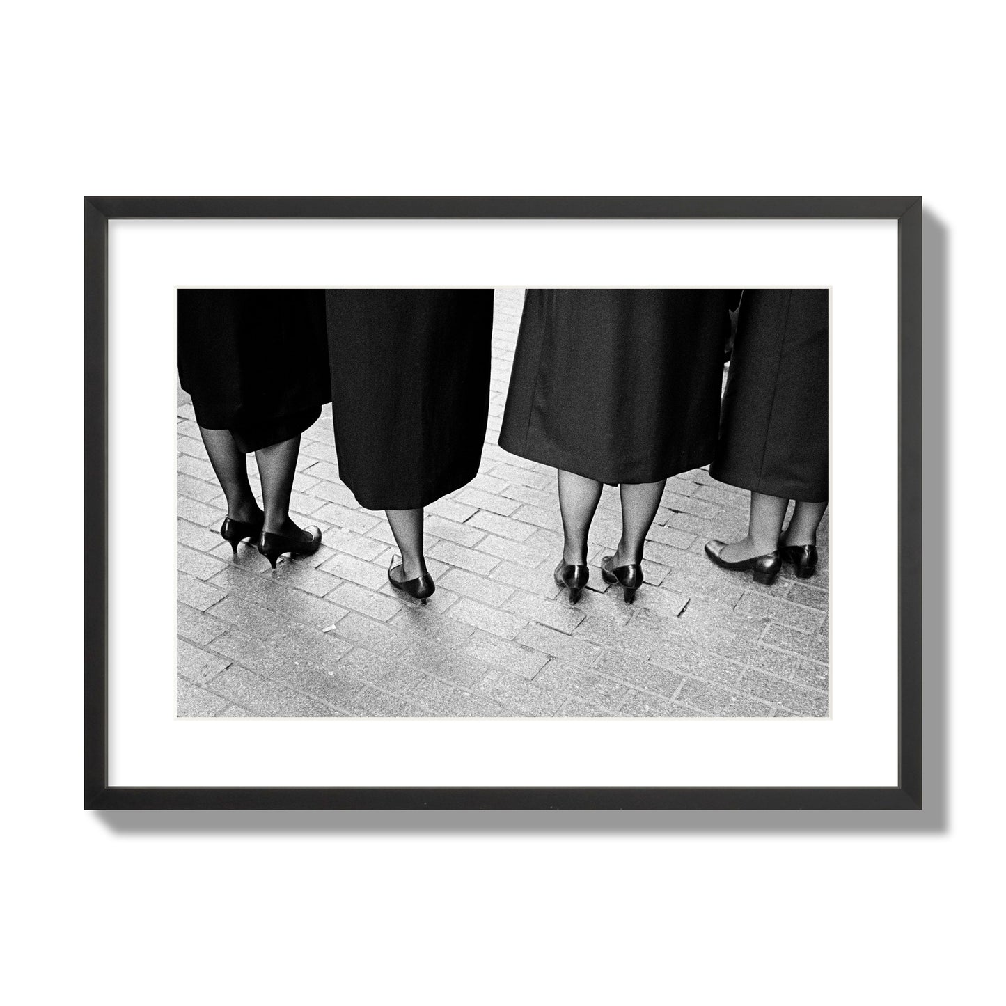 Fine art photograph depicting four women’s feet in high heels standing on a brick-paved street. One woman is notably missing a leg, adding a striking element to the composition, archival pigment print, large size, black frame