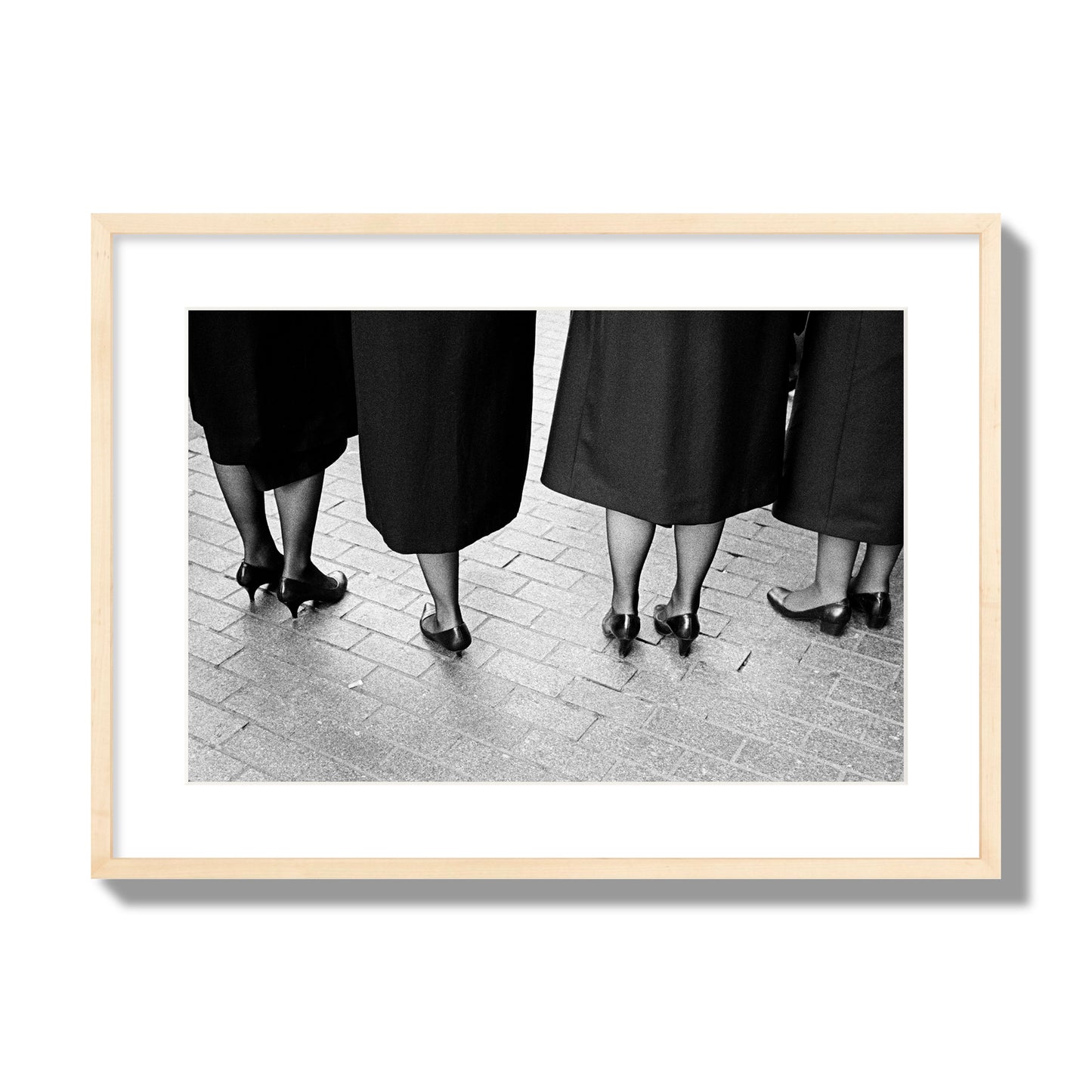 Fine art photograph depicting four women’s feet in high heels standing on a brick-paved street. One woman is notably missing a leg, adding a striking element to the composition, archival pigment print, large size, natural wood frame