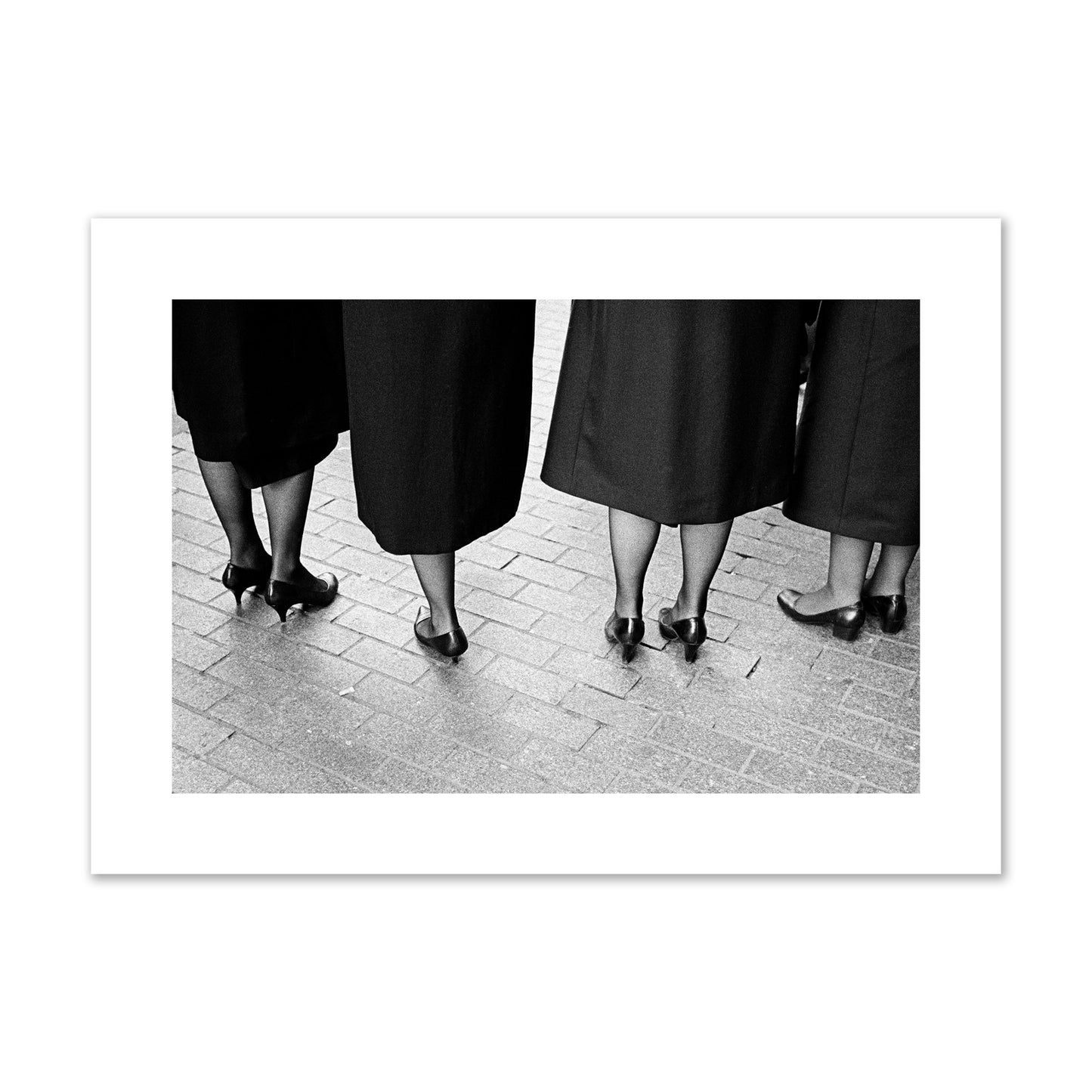 Fine art photograph depicting four women’s feet in high heels standing on a brick-paved street. One woman is notably missing a leg, adding a striking element to the composition, archival pigment print, large size, unframed.