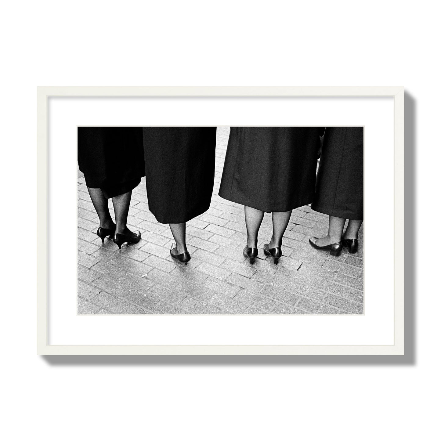 Fine art photograph depicting four women’s feet in high heels standing on a brick-paved street. One woman is notably missing a leg, adding a striking element to the composition, archival pigment print, large size, white frame