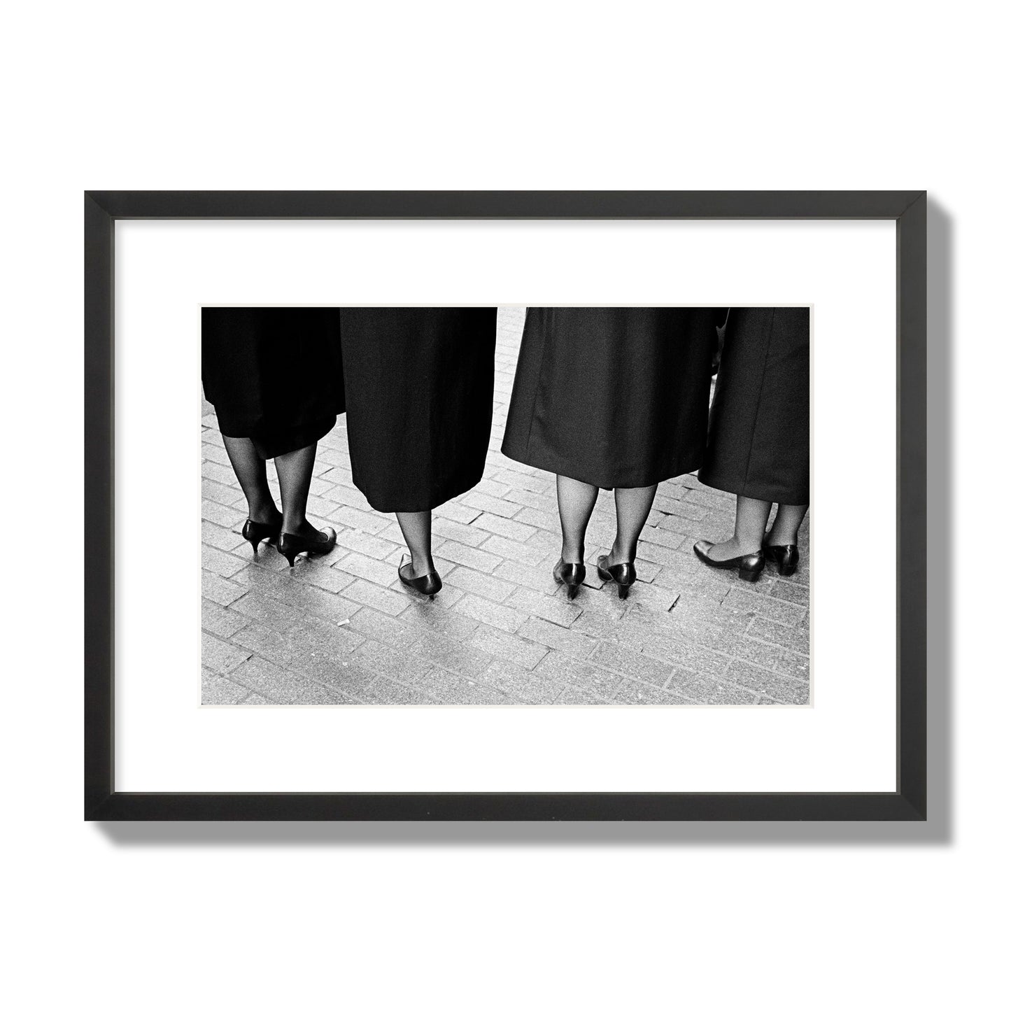 Fine art photograph depicting four women’s feet in high heels standing on a brick-paved street. One woman is notably missing a leg, adding a striking element to the composition, archival pigment print, medium size, black frame