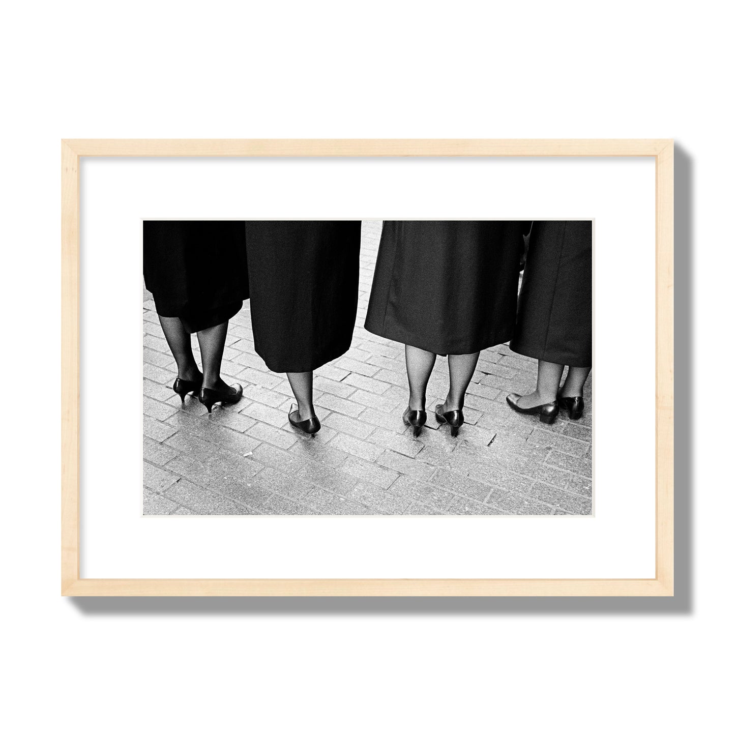 Fine art photograph depicting four women’s feet in high heels standing on a brick-paved street. One woman is notably missing a leg, adding a striking element to the composition, archival pigment print, medium size, natural wood frame