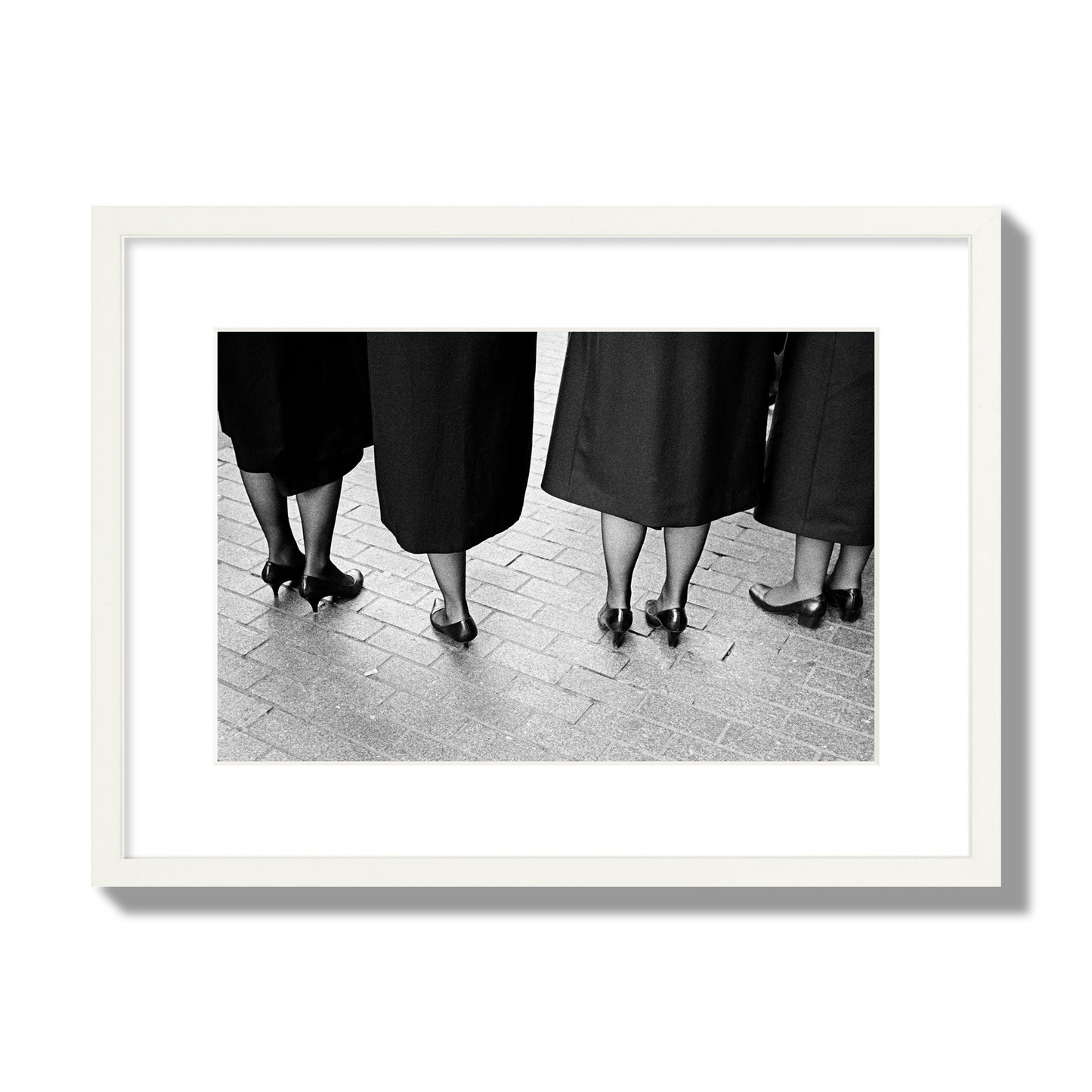 Fine art photograph depicting four women’s feet in high heels standing on a brick-paved street. One woman is notably missing a leg, adding a striking element to the composition, archival pigment print, medium size, white frame
