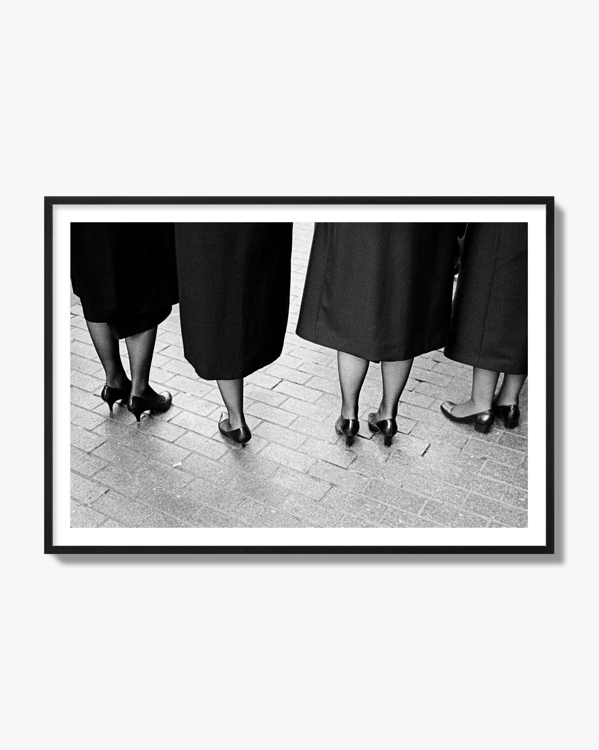 Fine art photograph depicting four women’s feet in high heels standing on a brick-paved street. One woman is notably missing a leg, adding a striking element to the composition, archival pigment print, black frame