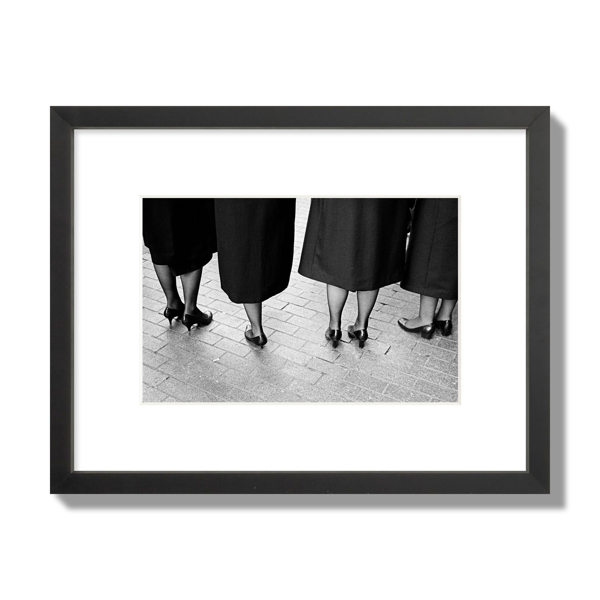 Fine art photograph depicting four women’s feet in high heels standing on a brick-paved street. One woman is notably missing a leg, adding a striking element to the composition, archival pigment print, small size, black frame