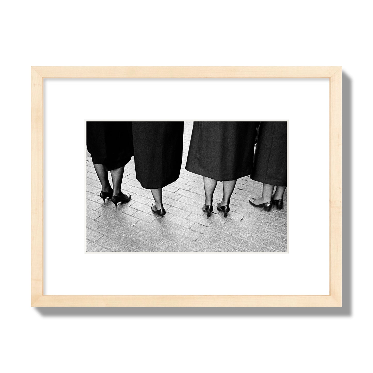 Fine art photograph depicting four women’s feet in high heels standing on a brick-paved street. One woman is notably missing a leg, adding a striking element to the composition, archival pigment print, small size, natural wood frame