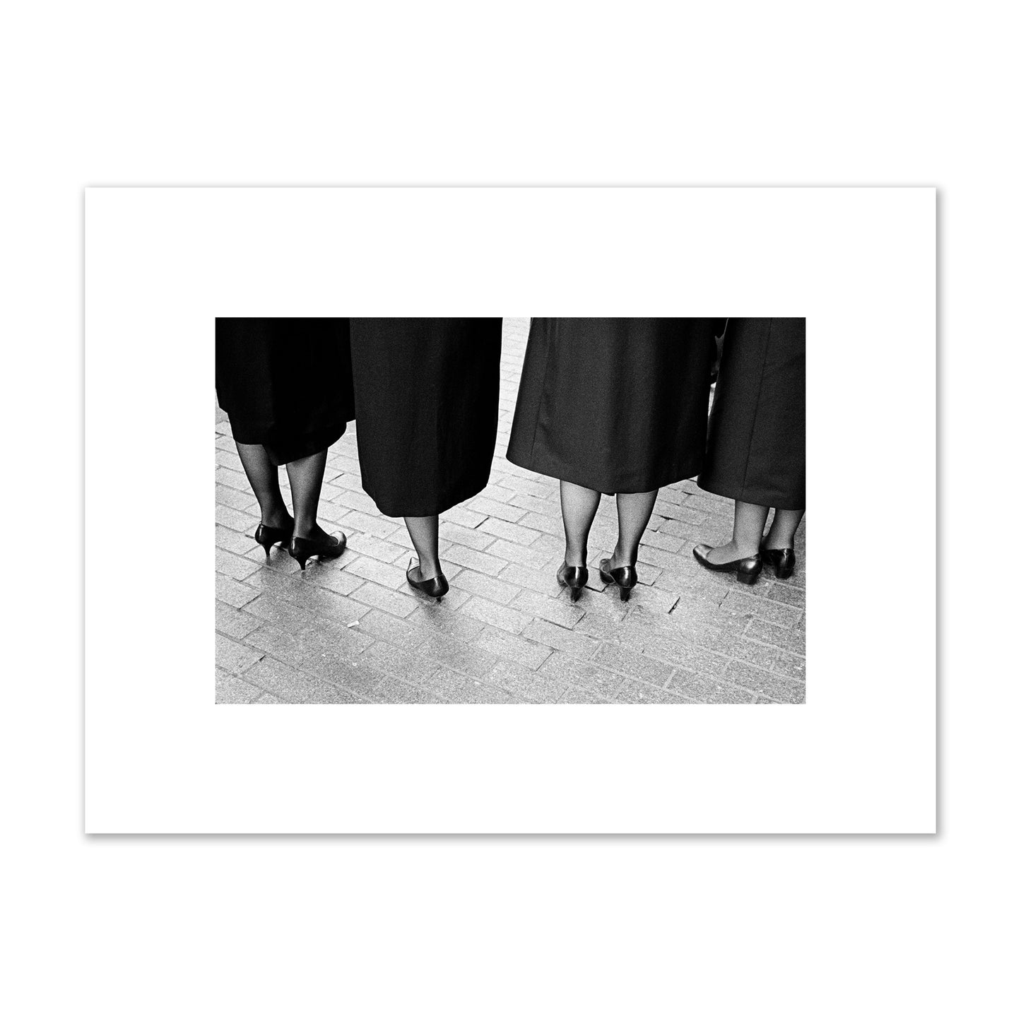 Fine art photograph depicting four women’s feet in high heels standing on a brick-paved street. One woman is notably missing a leg, adding a striking element to the composition, archival pigment print, small size, unframed.