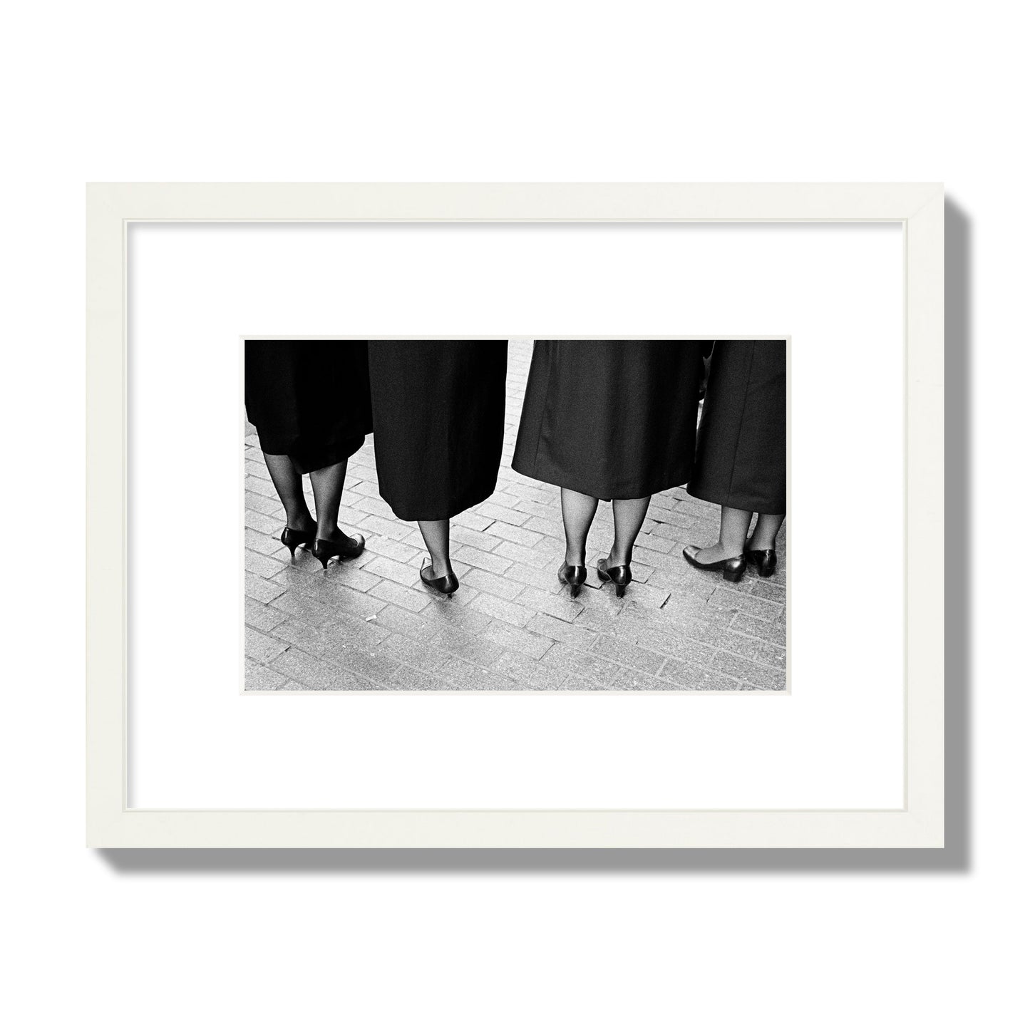Fine art photograph depicting four women’s feet in high heels standing on a brick-paved street. One woman is notably missing a leg, adding a striking element to the composition, archival pigment print, small size, white frame