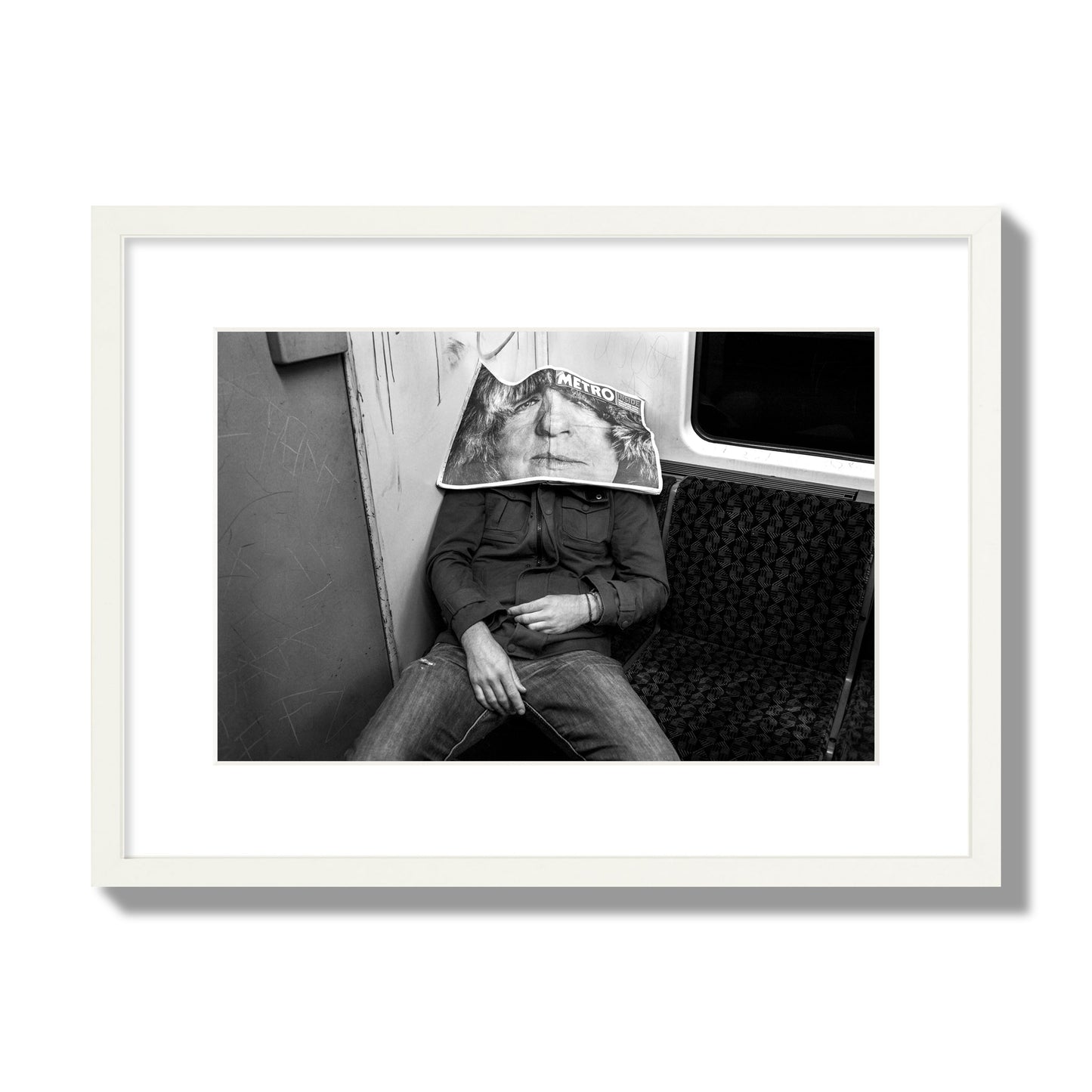 Fine art photograph of a man asleep on the subway with a newspaper covering their face, archival pigment print, medium size, white frame.