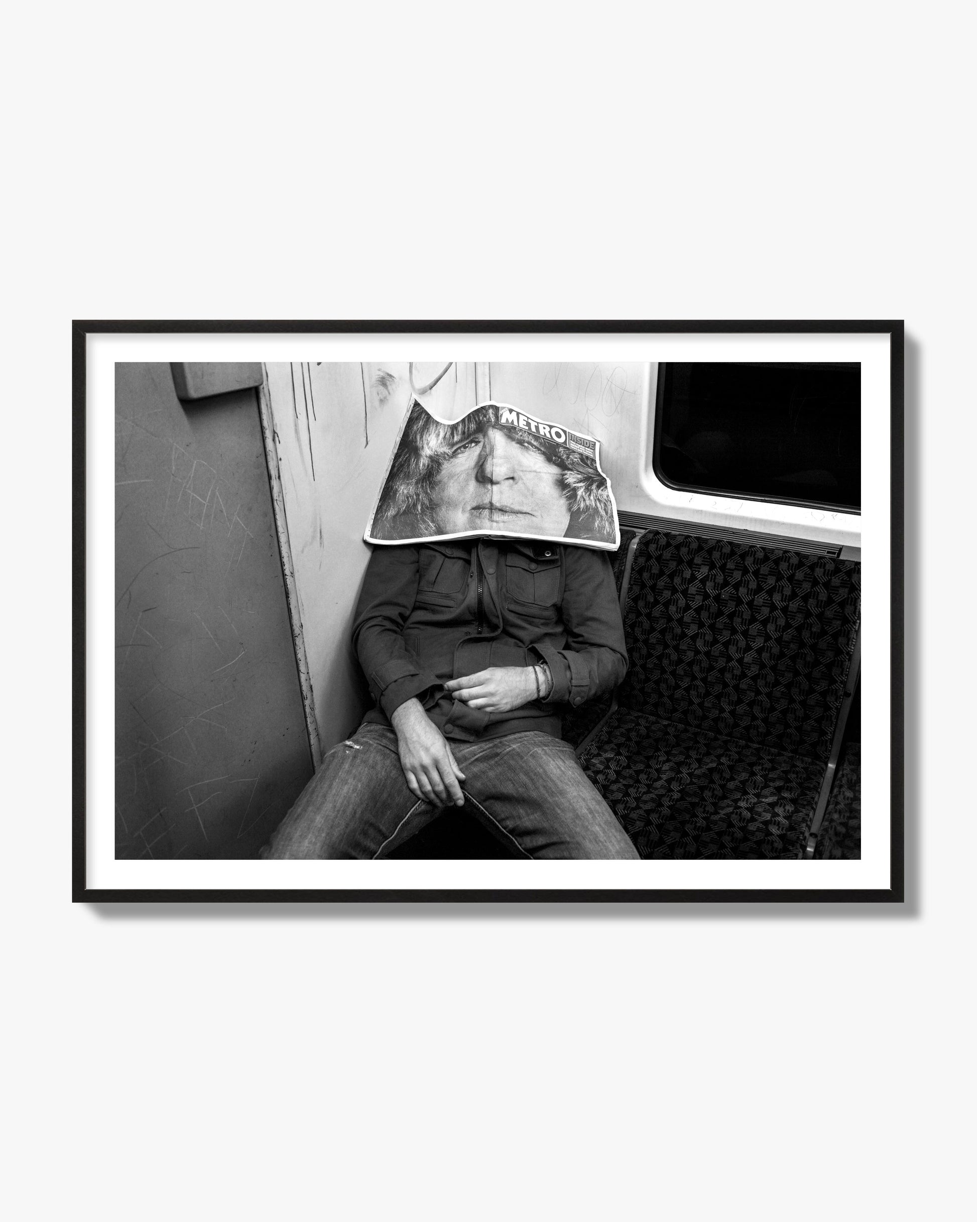 Fine art photograph of a man asleep on the subway with a newspaper covering their face, archival pigment print, black frame.