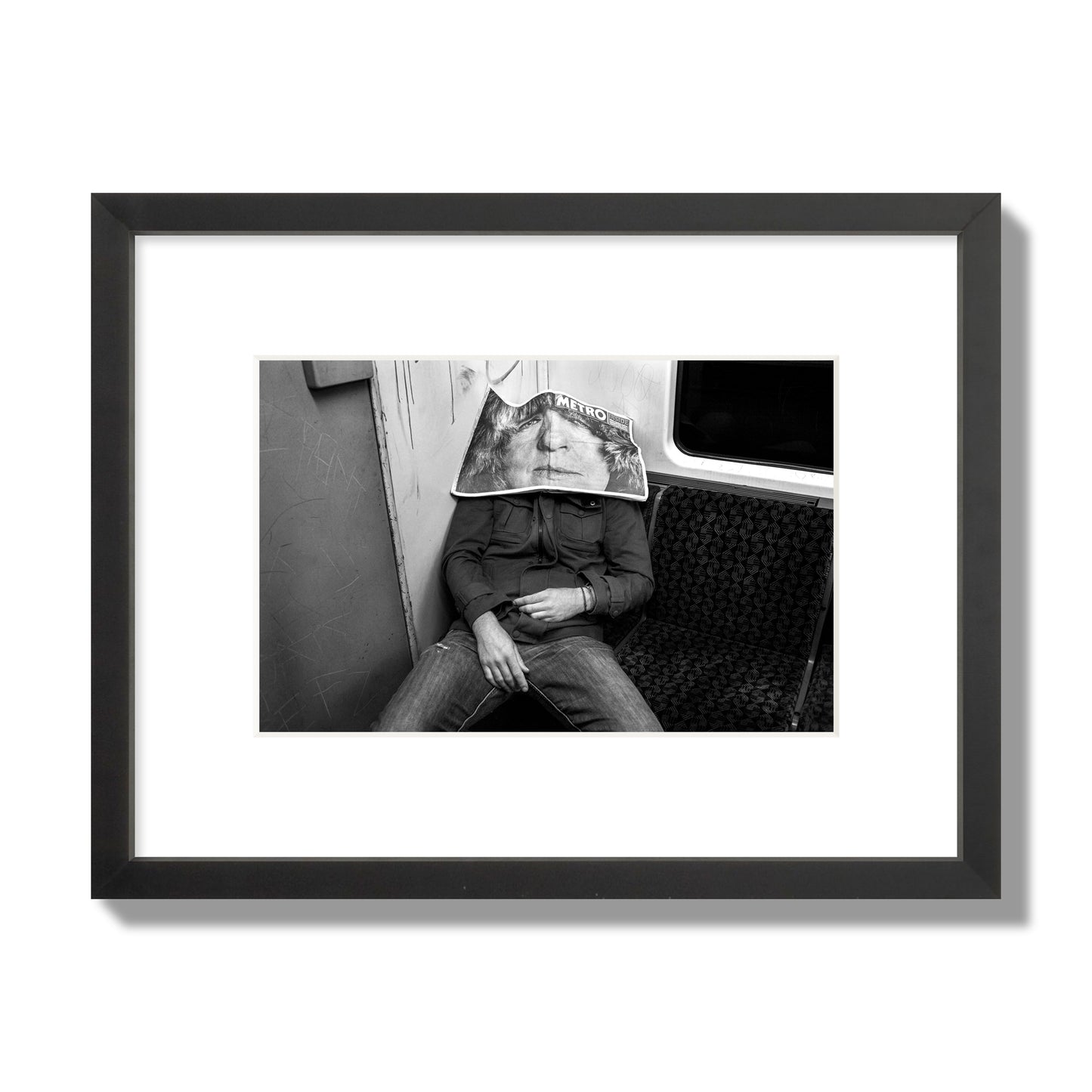 Fine art photograph of a man asleep on the subway with a newspaper covering their face, archival pigment print, small size, black frame.