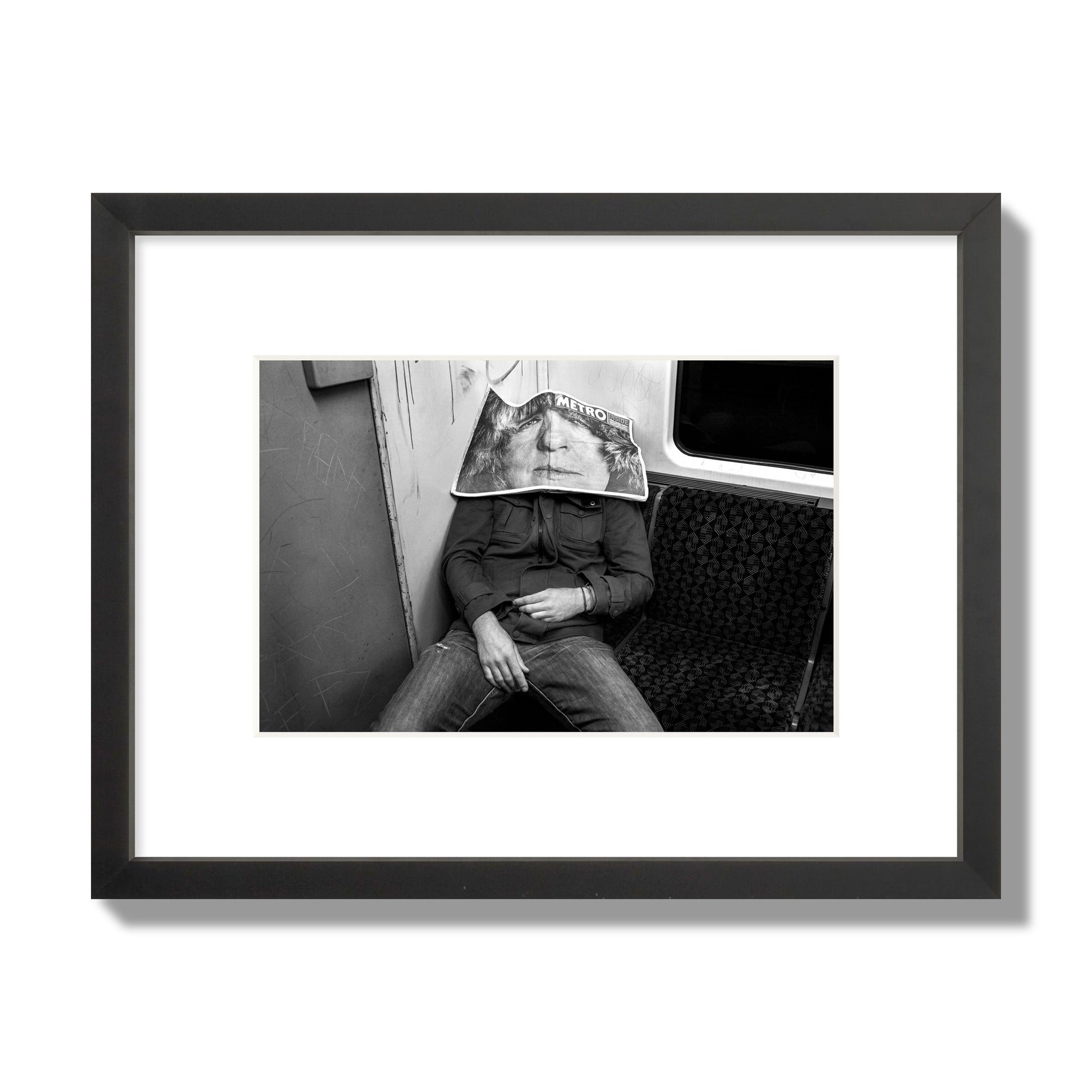 Fine art photograph of a man asleep on the subway with a newspaper covering their face, archival pigment print, small size, black frame.