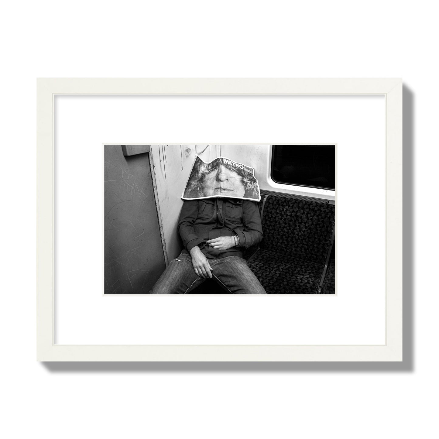 Fine art photograph of a man asleep on the subway with a newspaper covering their face, archival pigment print, small size, white frame.