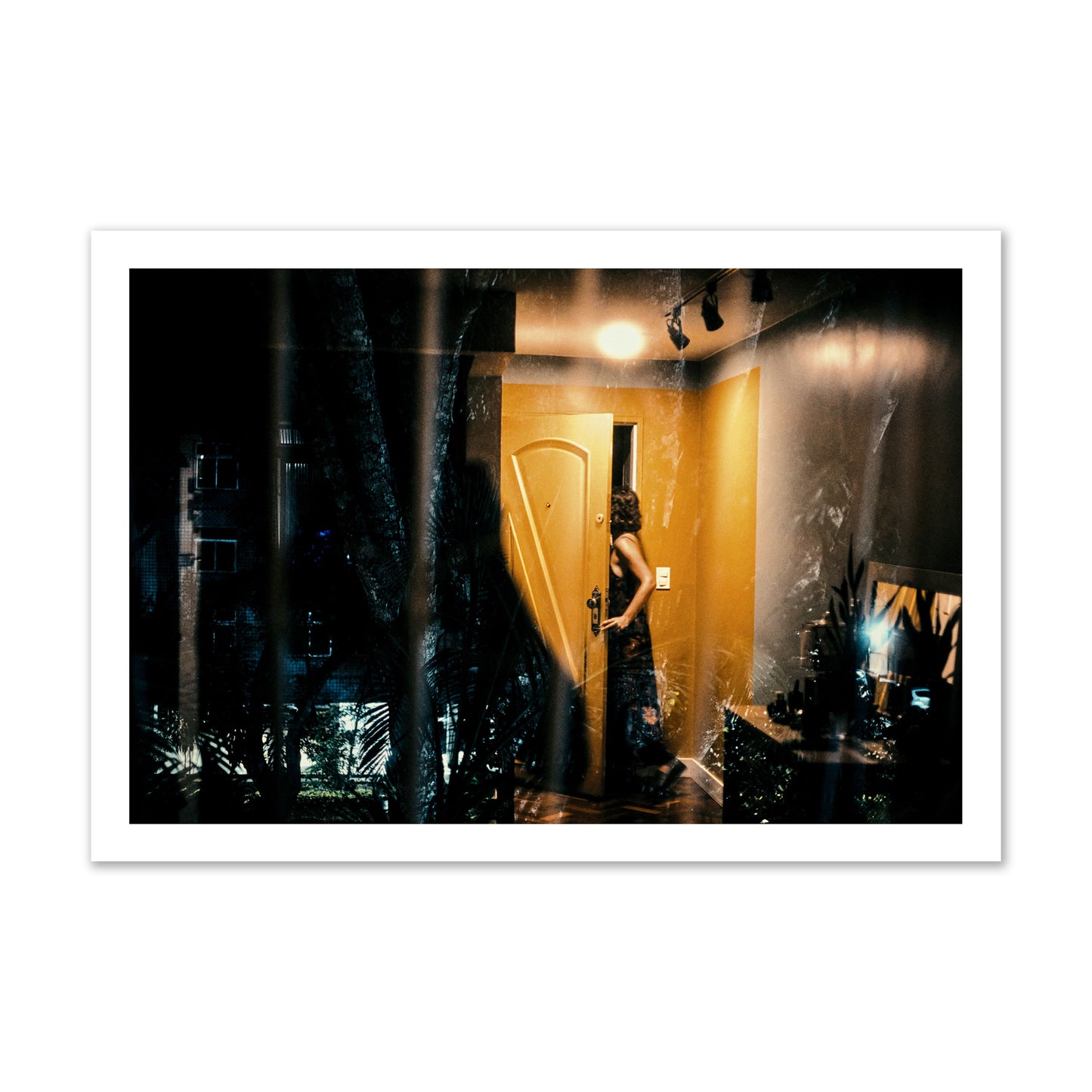 Fine art street photograph of a woman opening a bright yellow door to her living room, with reflections of the outside environment layered over the scene. Archival pigment print, gallery size, unframed.