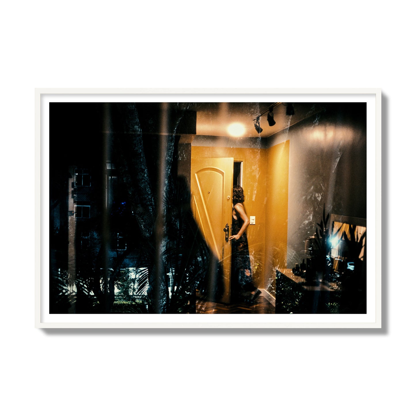 Fine art street photograph of a woman opening a bright yellow door to her living room, with reflections of the outside environment layered over the scene. Archival pigment print, gallery size, white frame.
