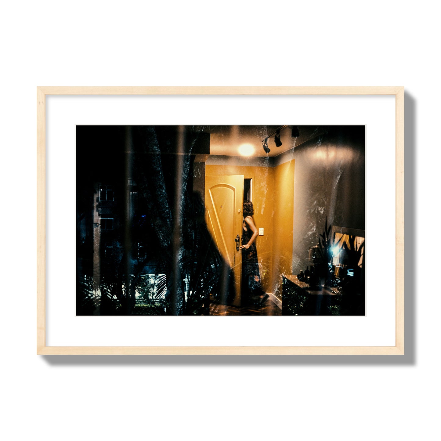 Fine art street photograph of a woman opening a bright yellow door to her living room, with reflections of the outside environment layered over the scene. Archival pigment print, large size, natural wood frame.