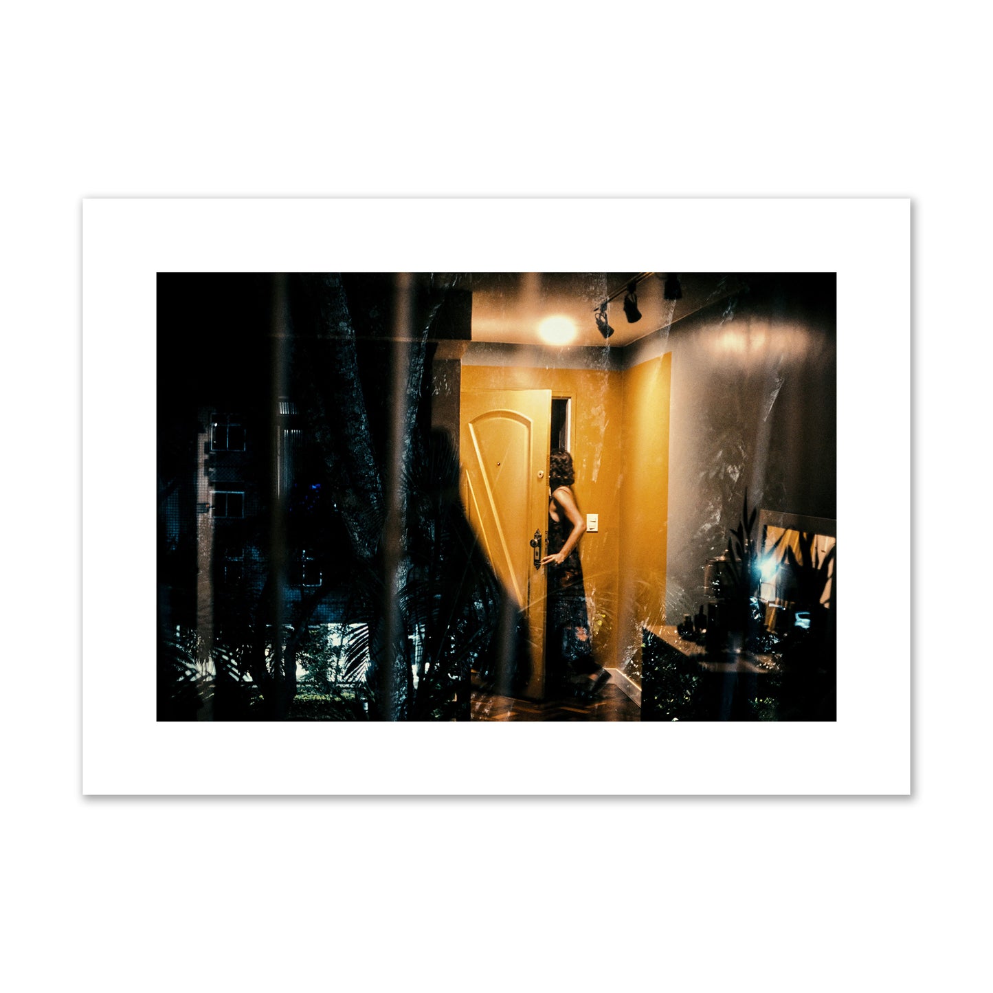 Fine art street photograph of a woman opening a bright yellow door to her living room, with reflections of the outside environment layered over the scene. Archival pigment print, large size, unframed.