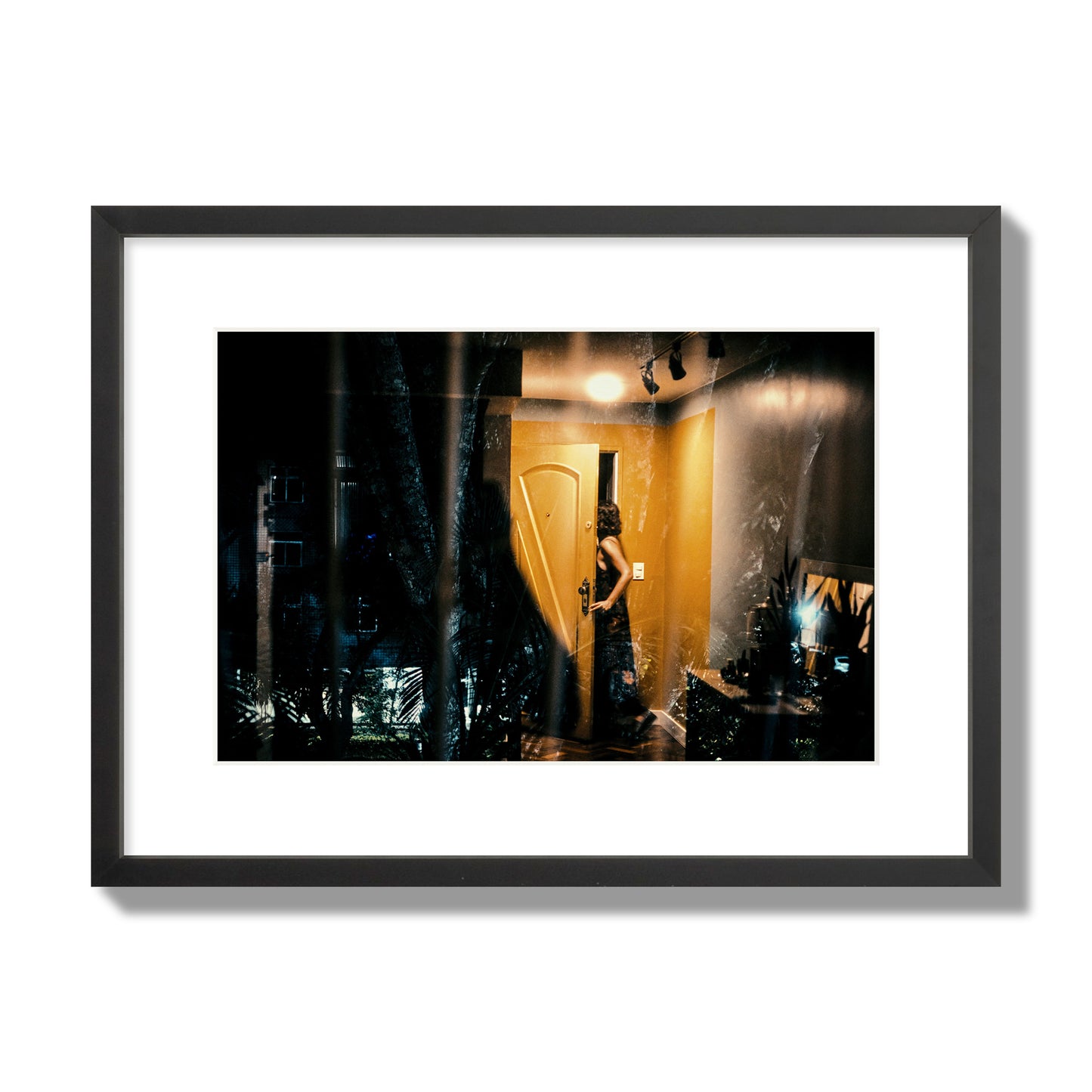 Fine art street photograph of a woman opening a bright yellow door to her living room, with reflections of the outside environment layered over the scene. Archival pigment print, medium size, black frame.