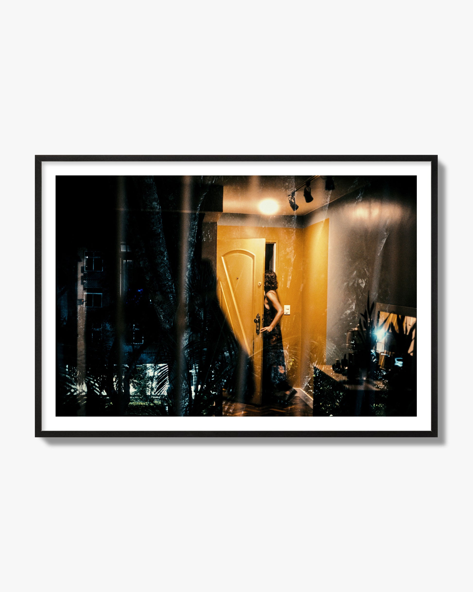 Fine art street photograph of a woman opening a bright yellow door to her living room, with reflections of the outside environment layered over the scene. Archival pigment print, black frame.