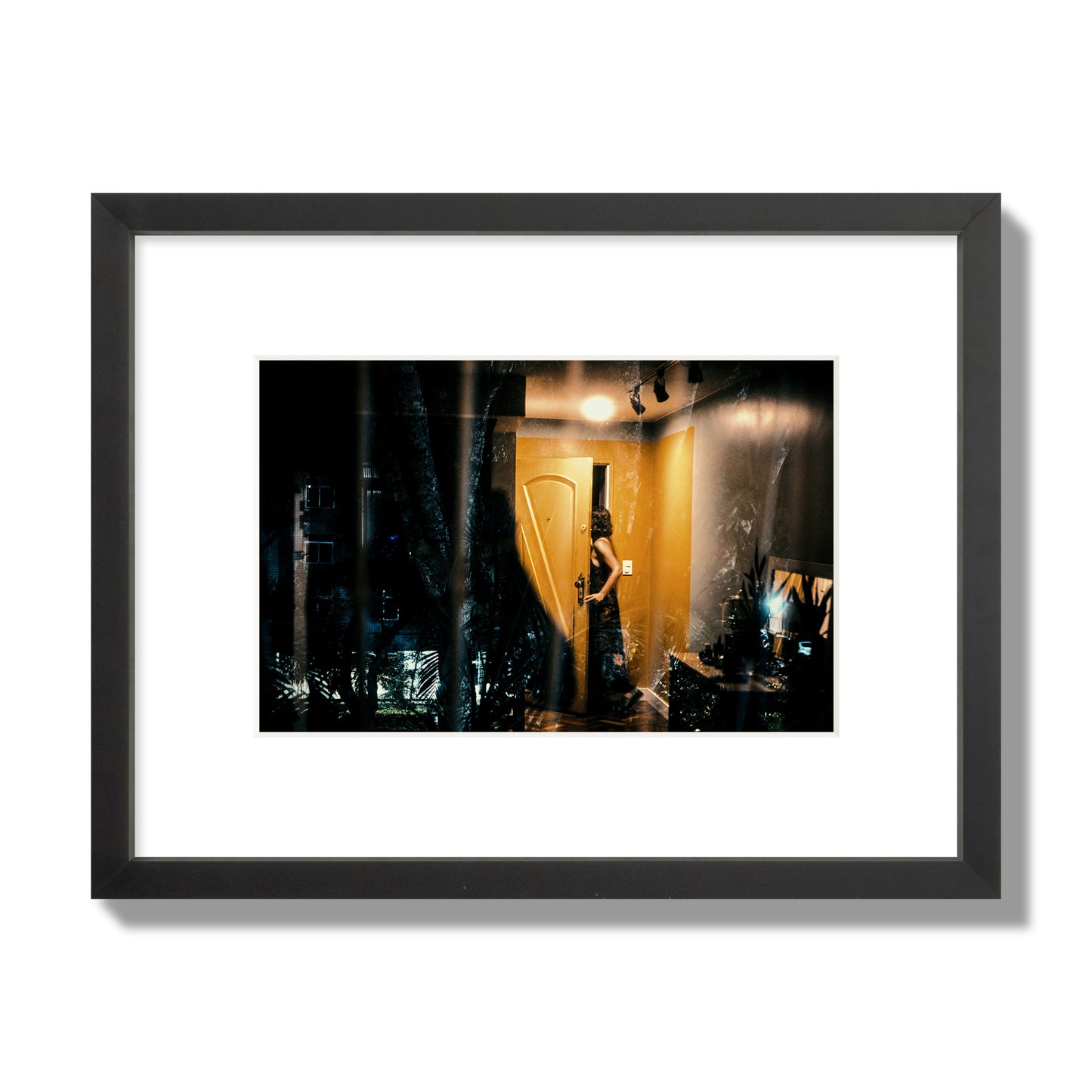 Fine art street photograph of a woman opening a bright yellow door to her living room, with reflections of the outside environment layered over the scene. Archival pigment print, small size, black frame.
