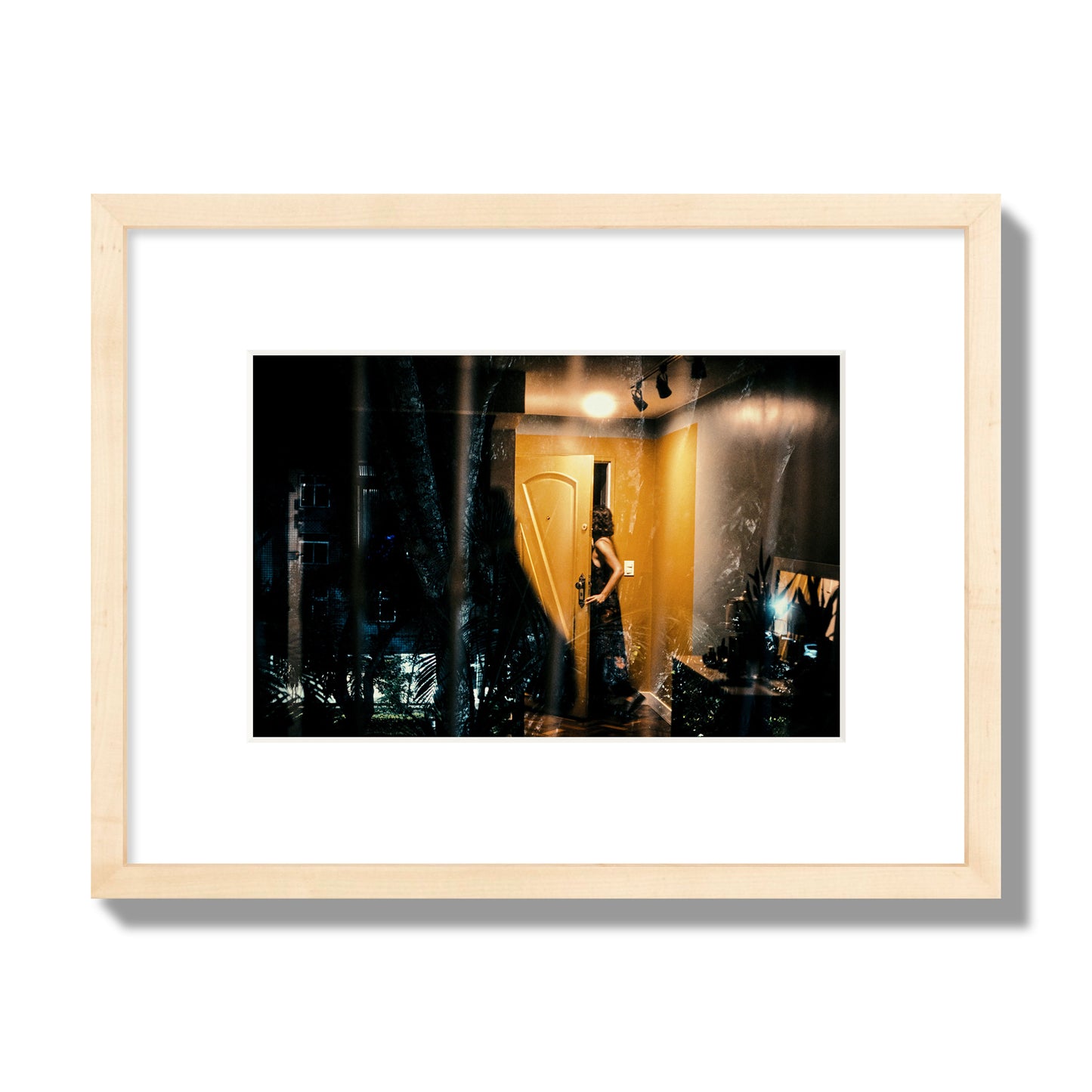 Fine art street photograph of a woman opening a bright yellow door to her living room, with reflections of the outside environment layered over the scene. Archival pigment print, small size, natural wood frame.