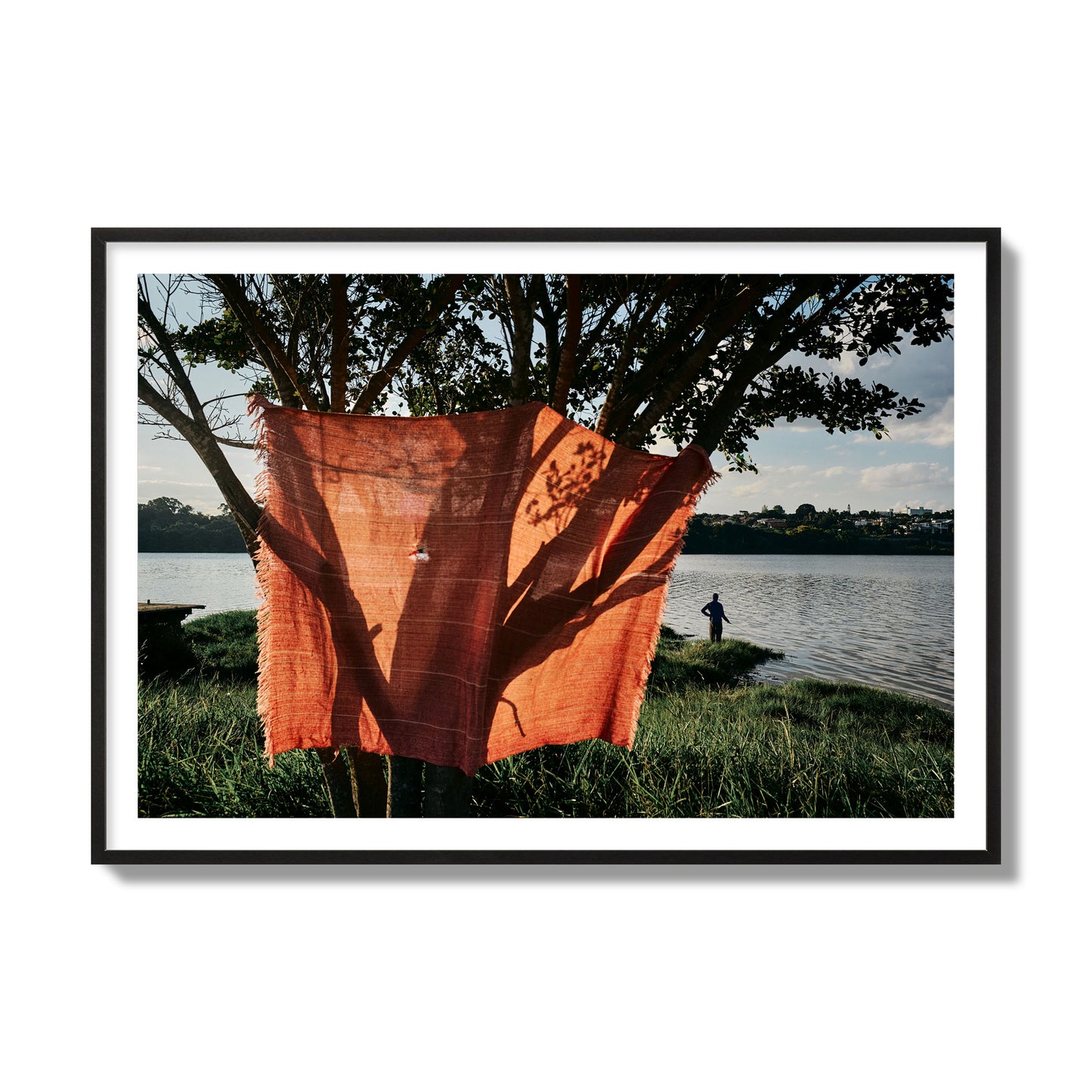 Fine art street photograph of a vibrant orange cloth hanging from a tree, with a tranquil lake and a fisherman in the background. Archival pigment print, collector size, black frame.