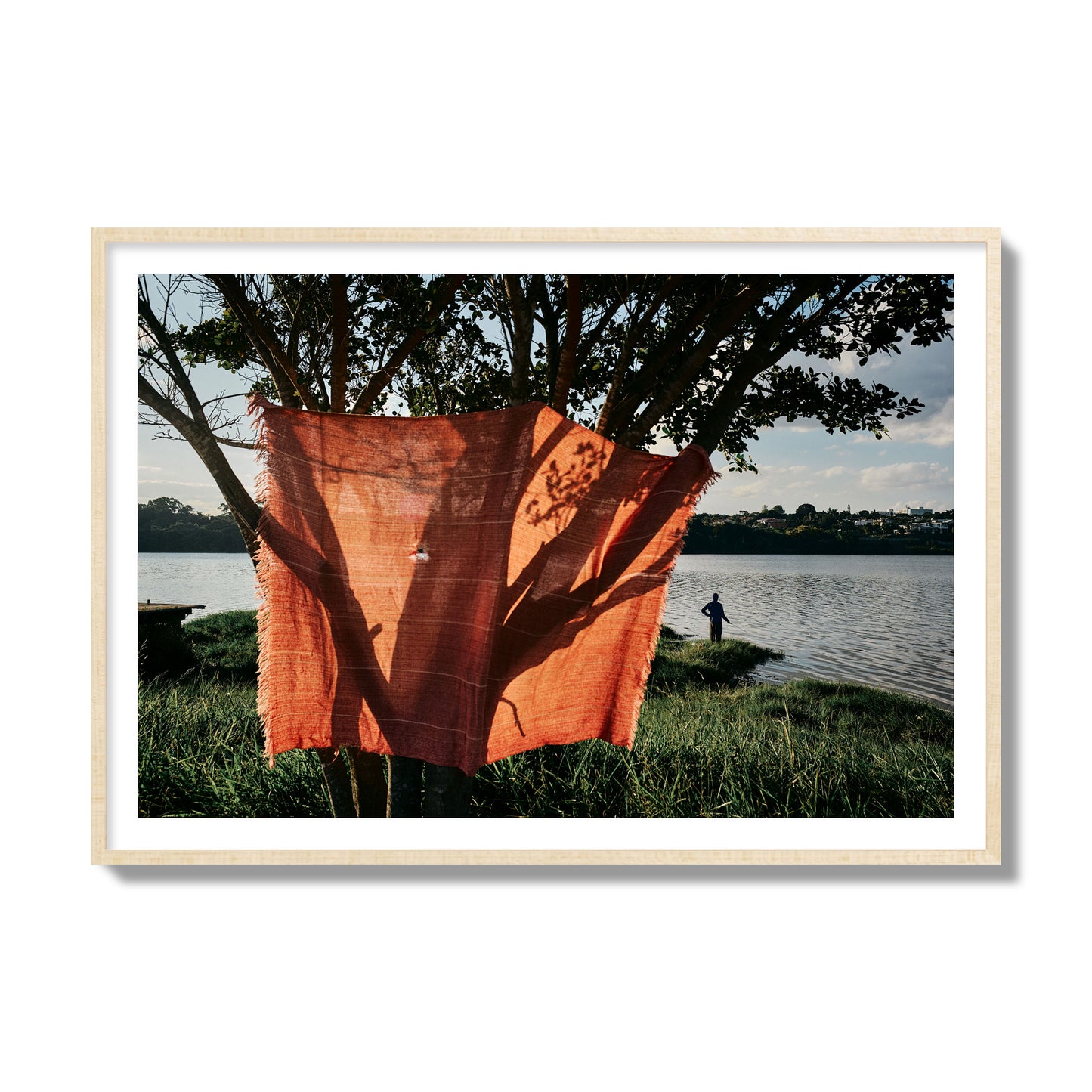 Fine art street photograph of a vibrant orange cloth hanging from a tree, with a tranquil lake and a fisherman in the background. Archival pigment print, collector size, natural wood frame.