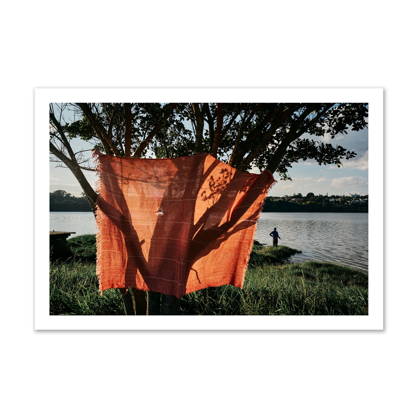 Fine art street photograph of a vibrant orange cloth hanging from a tree, with a tranquil lake and a fisherman in the background. Archival pigment print, gallery size, unframed.