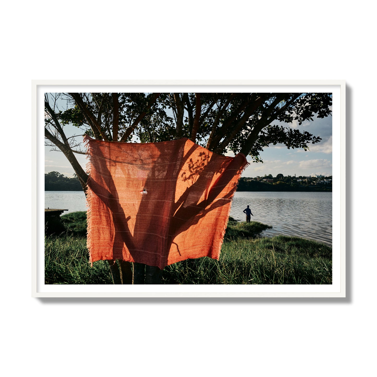 Fine art street photograph of a vibrant orange cloth hanging from a tree, with a tranquil lake and a fisherman in the background. Archival pigment print, gallery size, white frame.