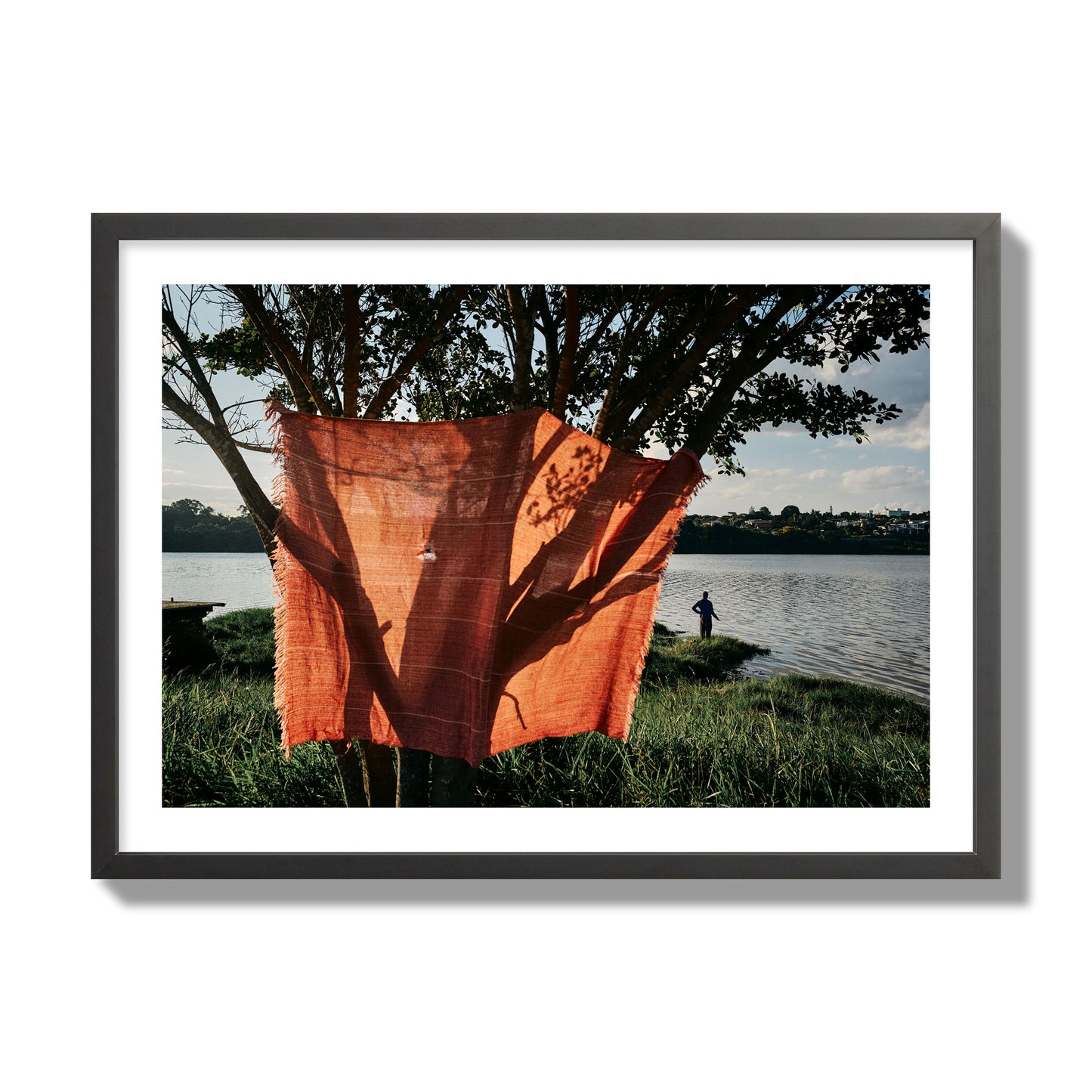 Fine art street photograph of a vibrant orange cloth hanging from a tree, with a tranquil lake and a fisherman in the background. Archival pigment print, giant size, black frame.