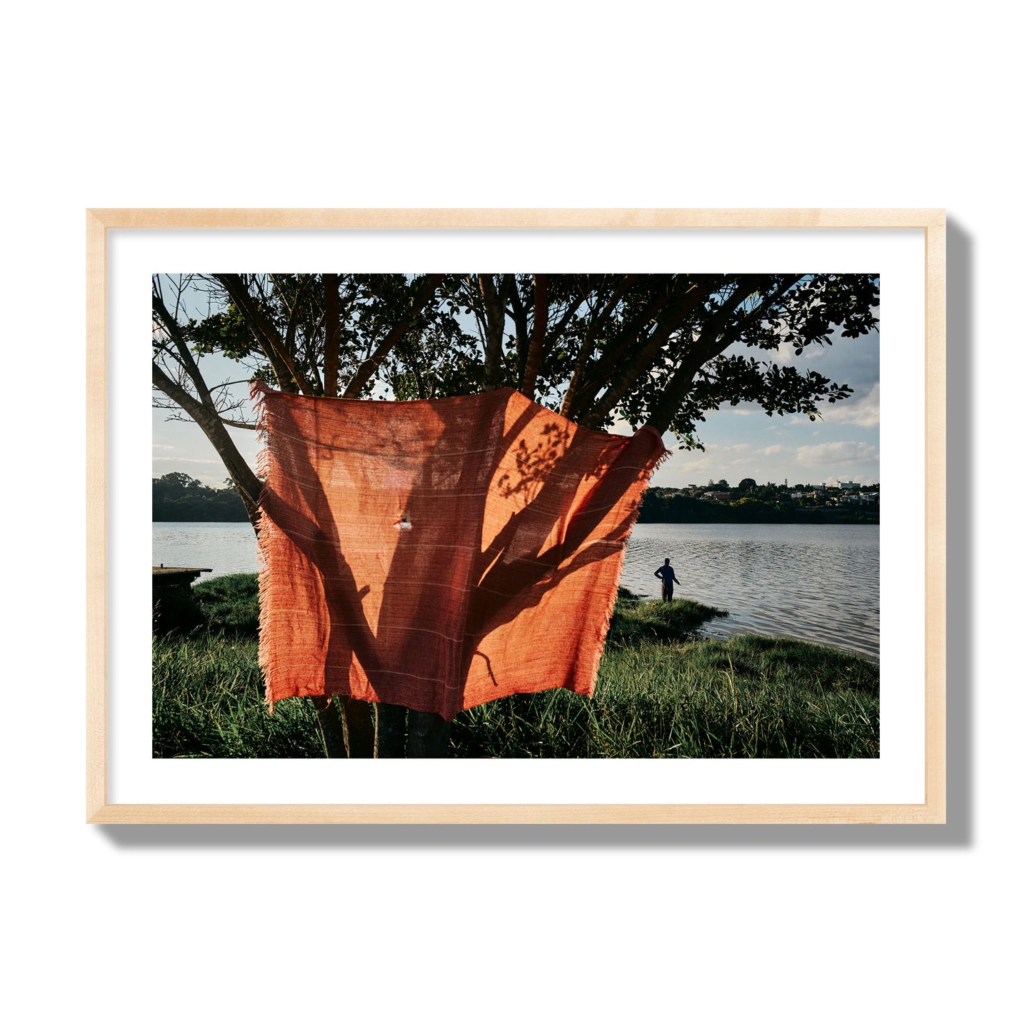 Fine art street photograph of a vibrant orange cloth hanging from a tree, with a tranquil lake and a fisherman in the background. Archival pigment print, giant size, natural wood frame.