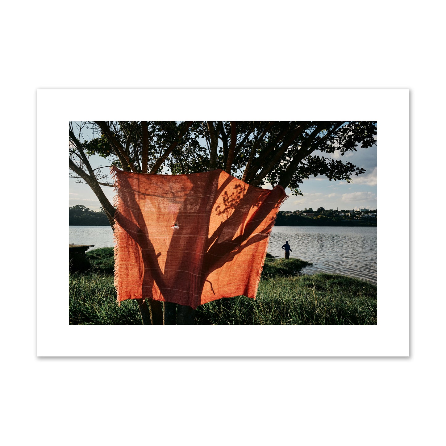 Fine art street photograph of a vibrant orange cloth hanging from a tree, with a tranquil lake and a fisherman in the background. Archival pigment print, giant size, unframed.