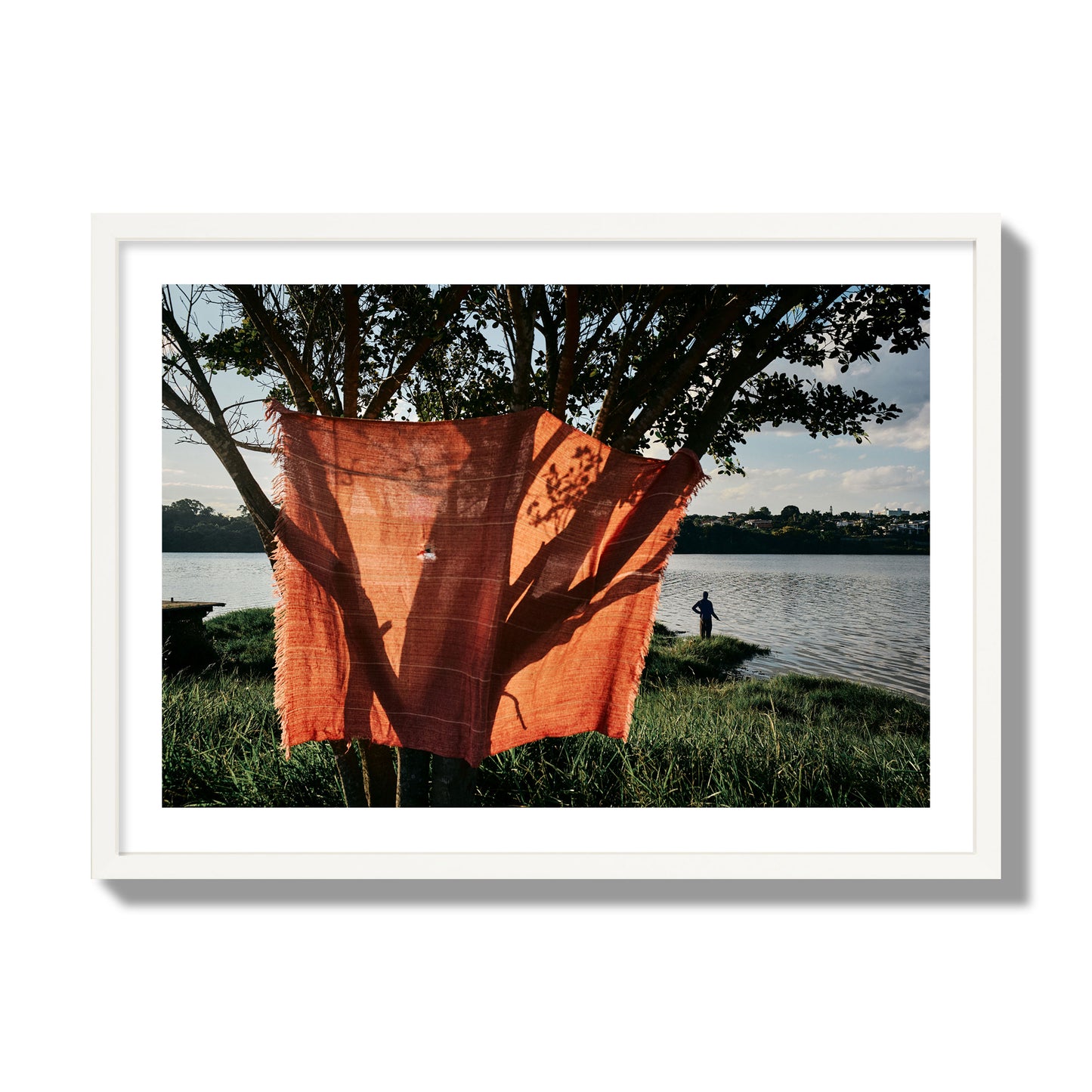Fine art street photograph of a vibrant orange cloth hanging from a tree, with a tranquil lake and a fisherman in the background. Archival pigment print, giant size, white frame.
