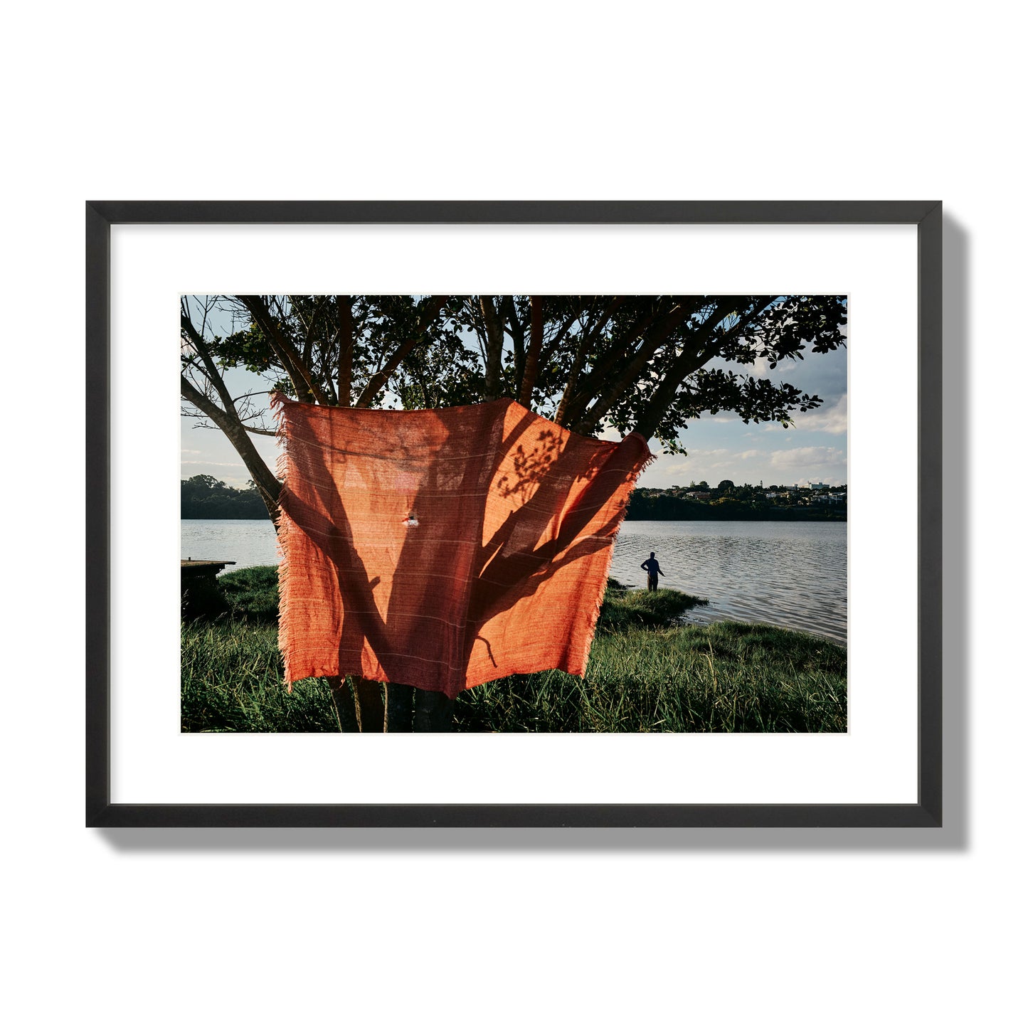 Fine art street photograph of a vibrant orange cloth hanging from a tree, with a tranquil lake and a fisherman in the background. Archival pigment print, large size, black frame.