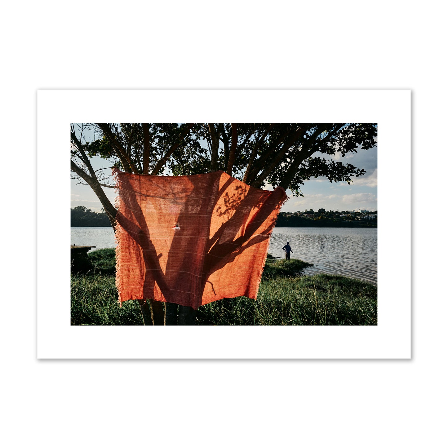Fine art street photograph of a vibrant orange cloth hanging from a tree, with a tranquil lake and a fisherman in the background. Archival pigment print, large size, unframed.