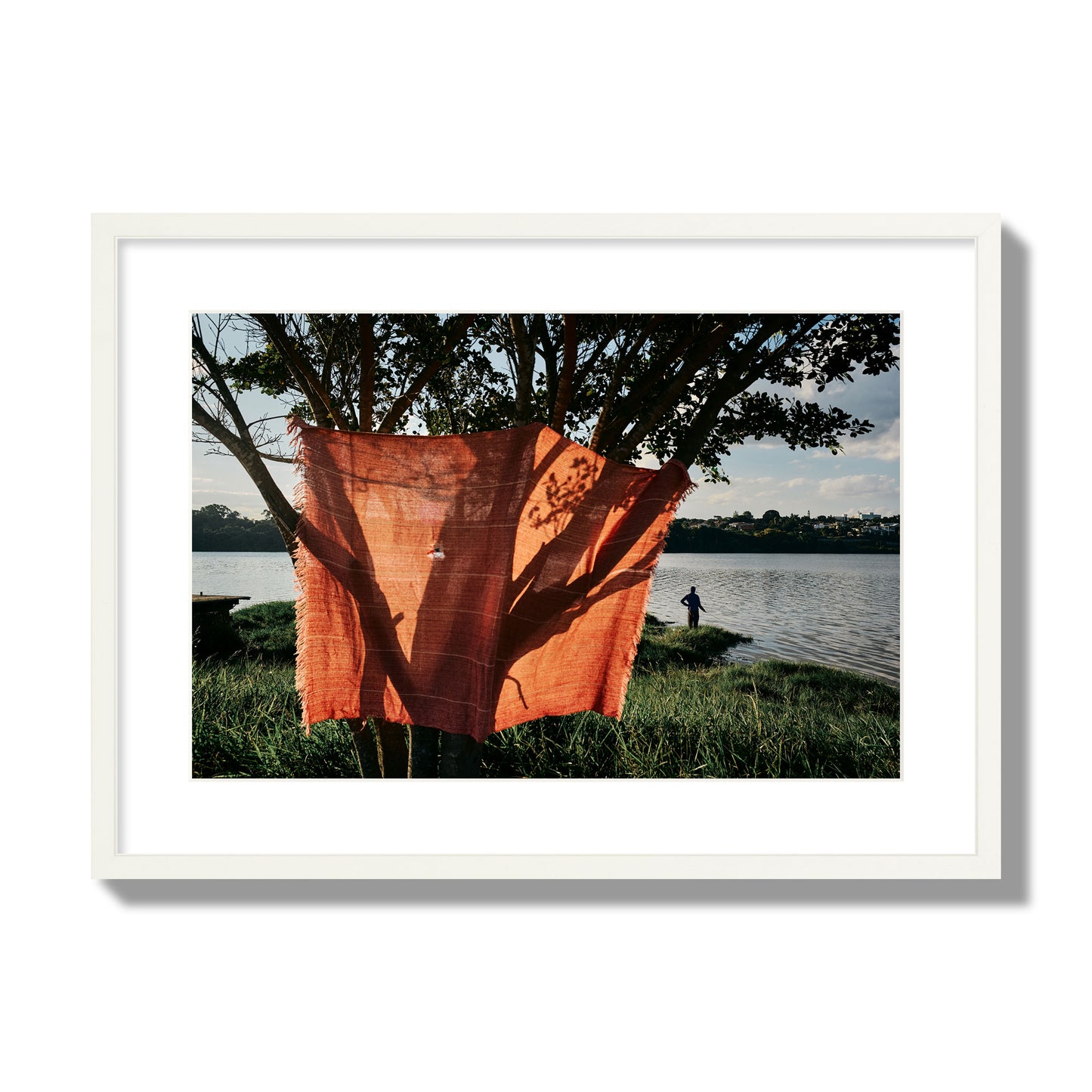 Fine art street photograph of a vibrant orange cloth hanging from a tree, with a tranquil lake and a fisherman in the background. Archival pigment print, large size, white frame.