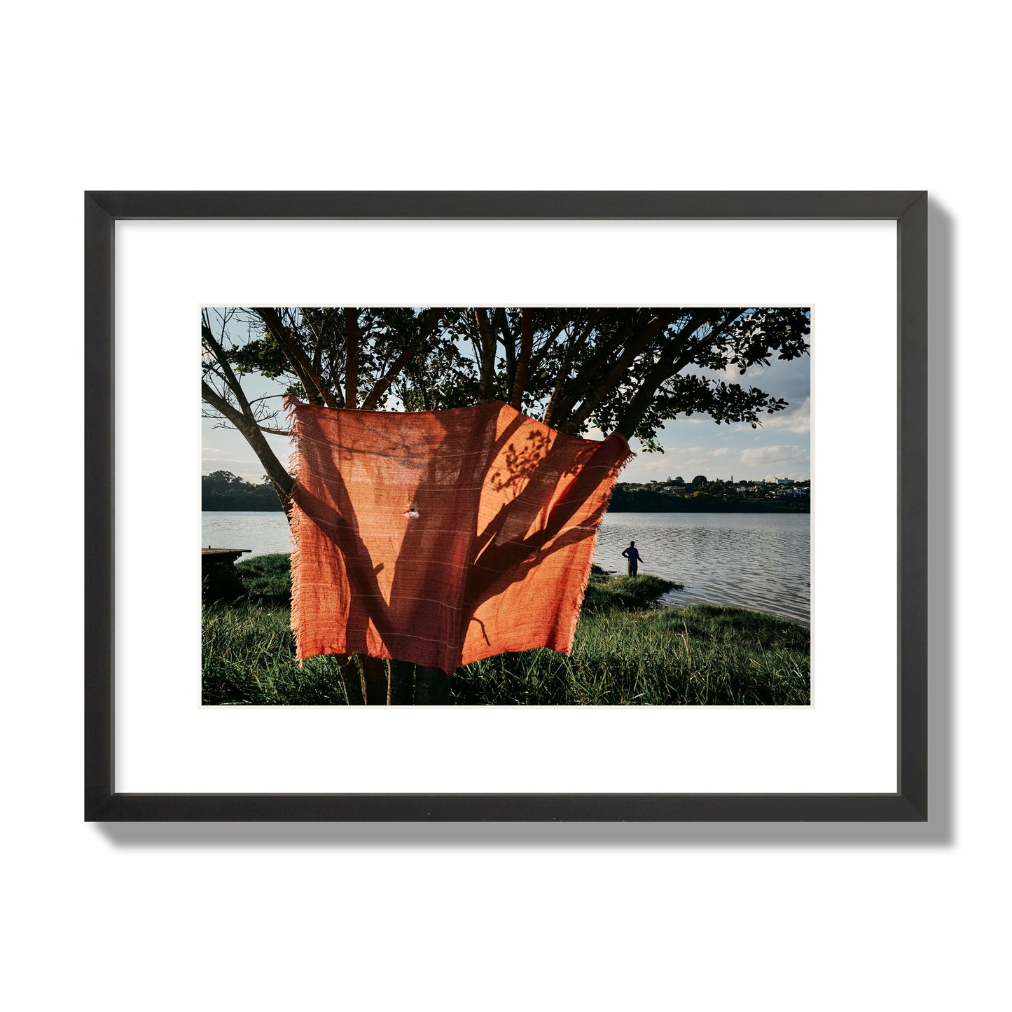 Fine art street photograph of a vibrant orange cloth hanging from a tree, with a tranquil lake and a fisherman in the background. Archival pigment print, medium size, black frame.