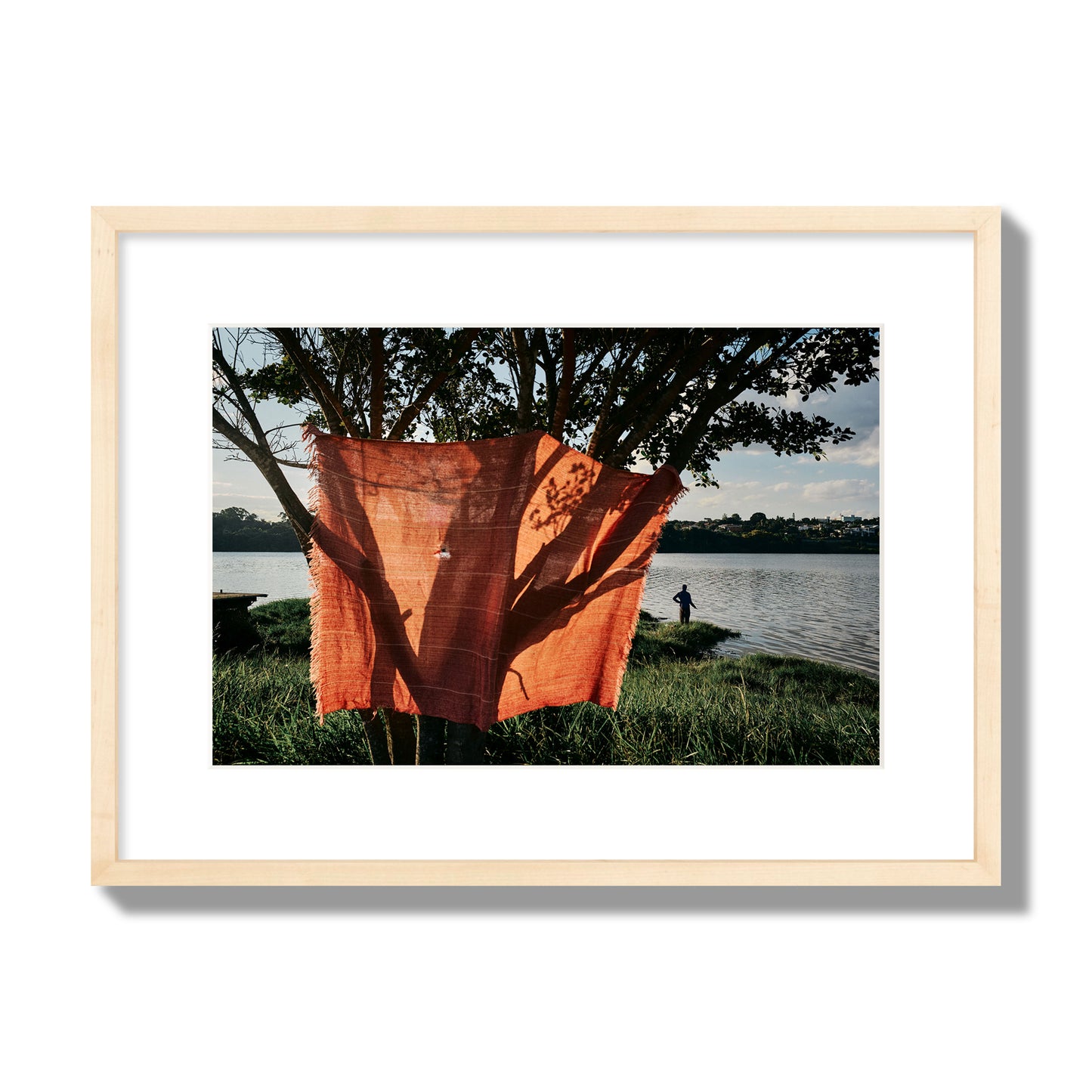 Fine art street photograph of a vibrant orange cloth hanging from a tree, with a tranquil lake and a fisherman in the background. Archival pigment print, medium size, natural wood frame.