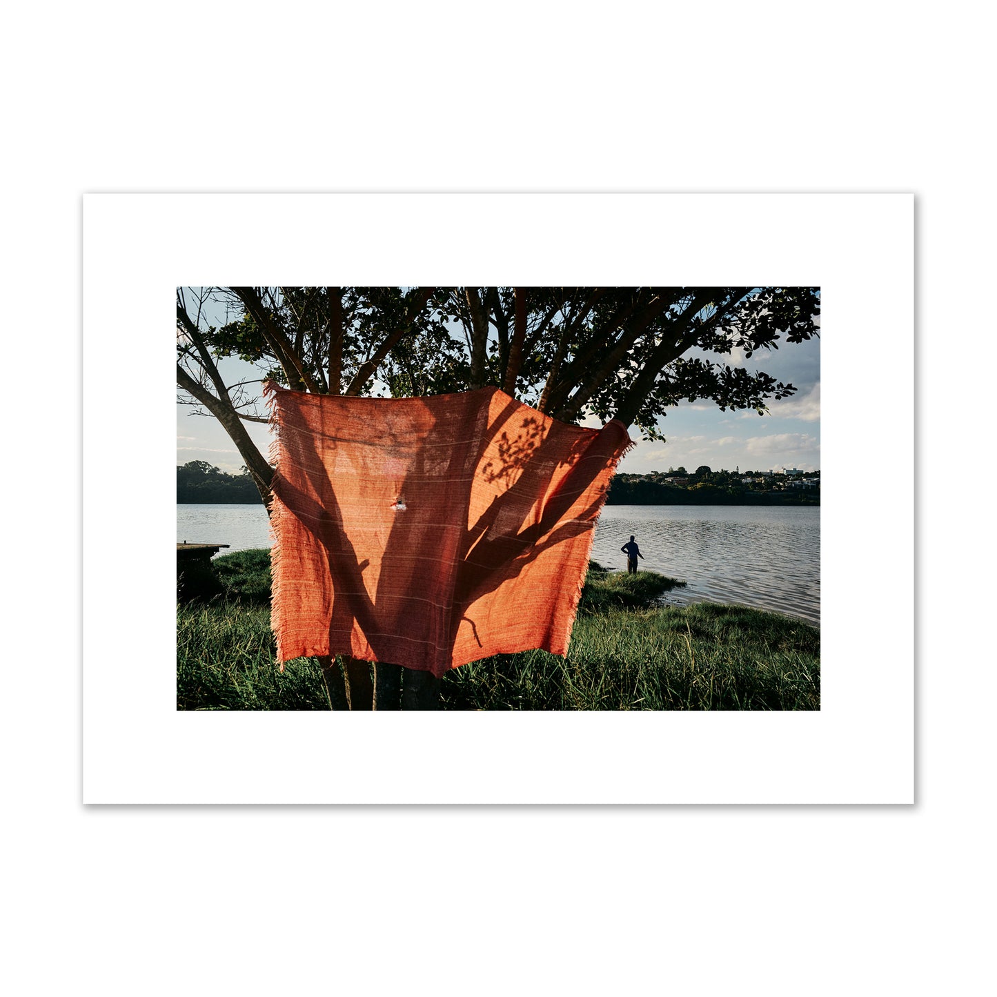 Fine art street photograph of a vibrant orange cloth hanging from a tree, with a tranquil lake and a fisherman in the background. Archival pigment print, medium size, unframed.