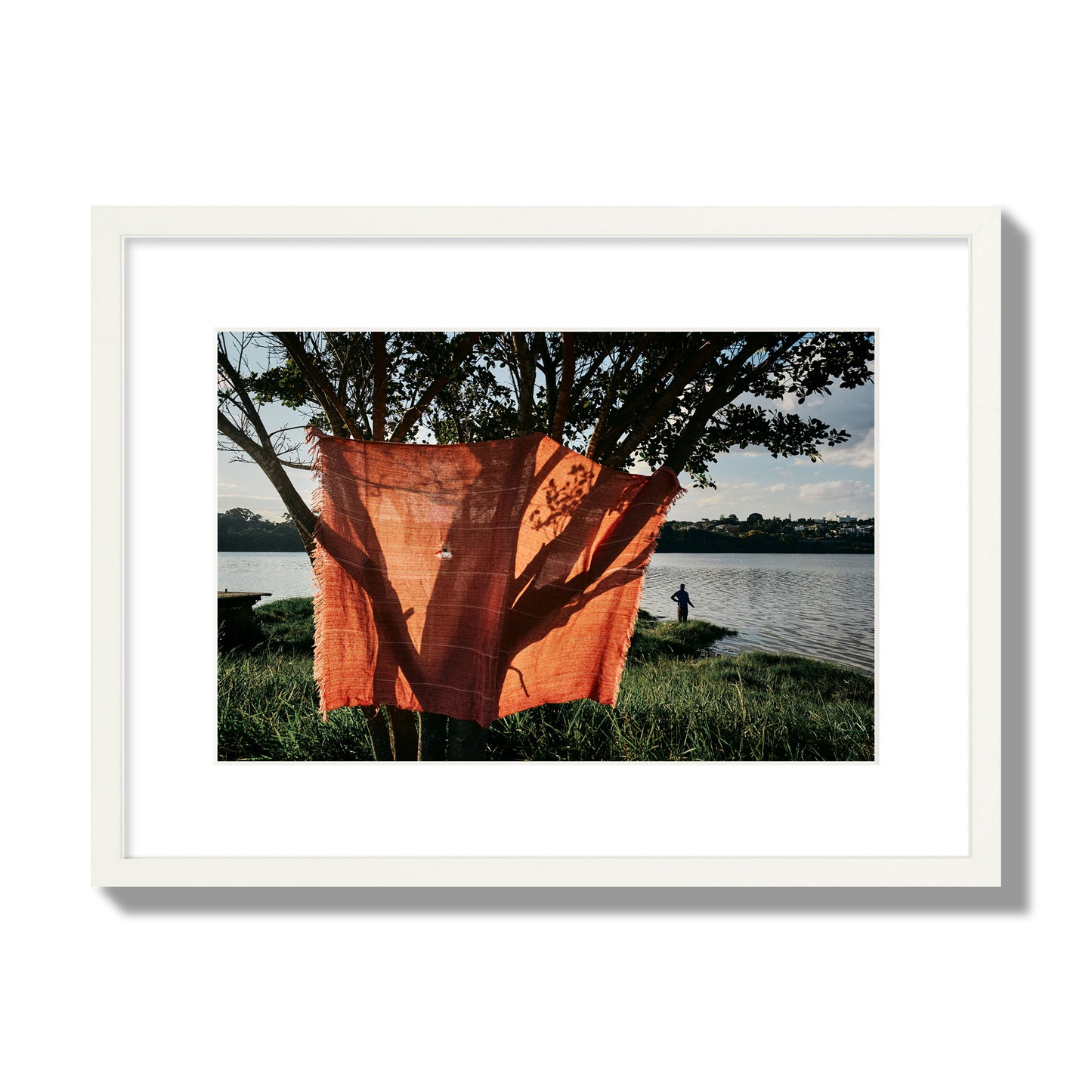 Fine art street photograph of a vibrant orange cloth hanging from a tree, with a tranquil lake and a fisherman in the background. Archival pigment print, medium size, white frame.